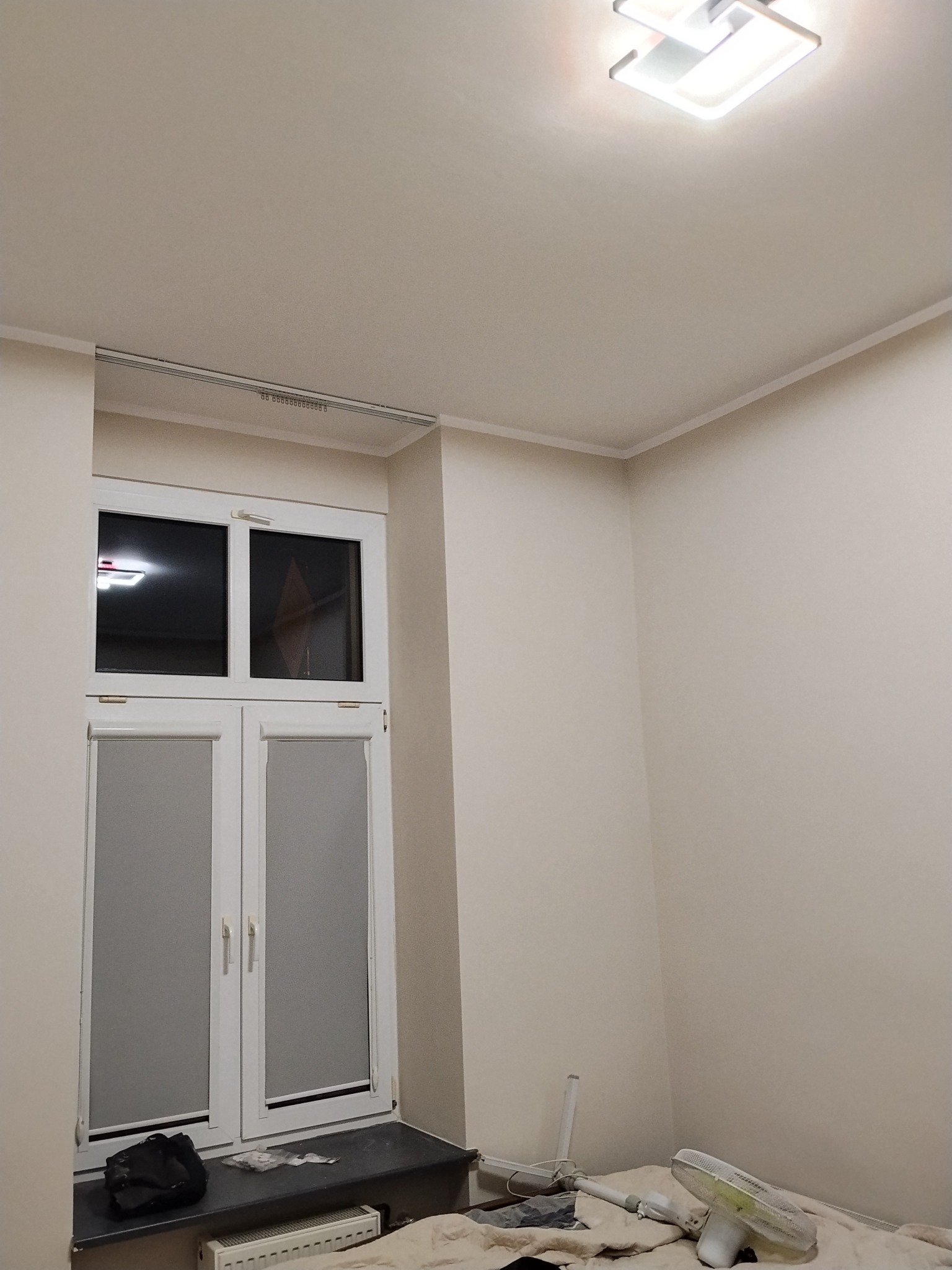 Pokój po remoncie: białe ściany, okno z roletami, grzejnik pod oknem, nowoczesna lampa sufitowa. Widoczny wentylator i torba na parapecie.