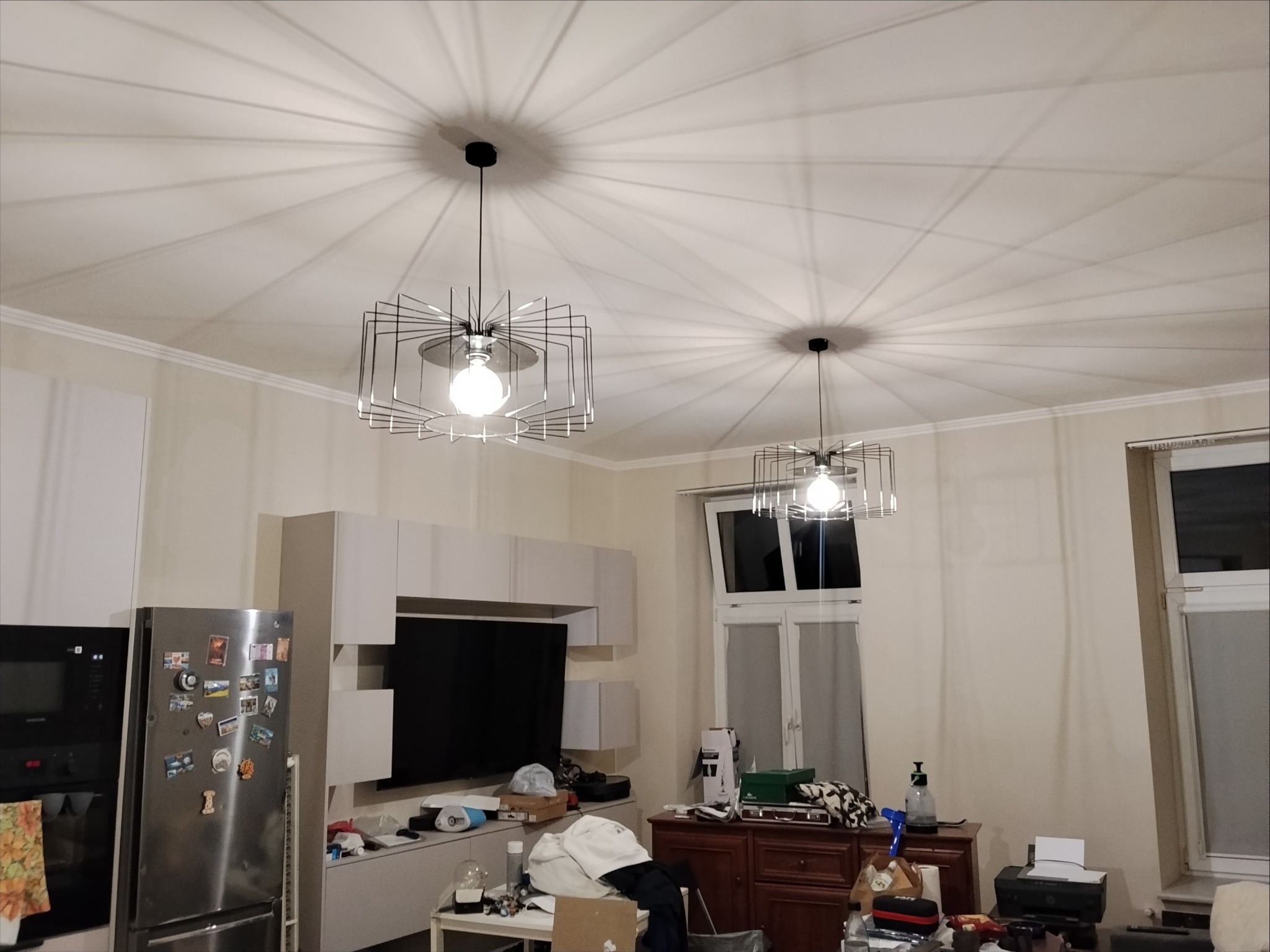 Dwa nowoczesne, geometryczne żyrandole z żarówkami kulistymi, rzucające promieniowe cienie na jasny sufit w pokoju z oknem i meblami.