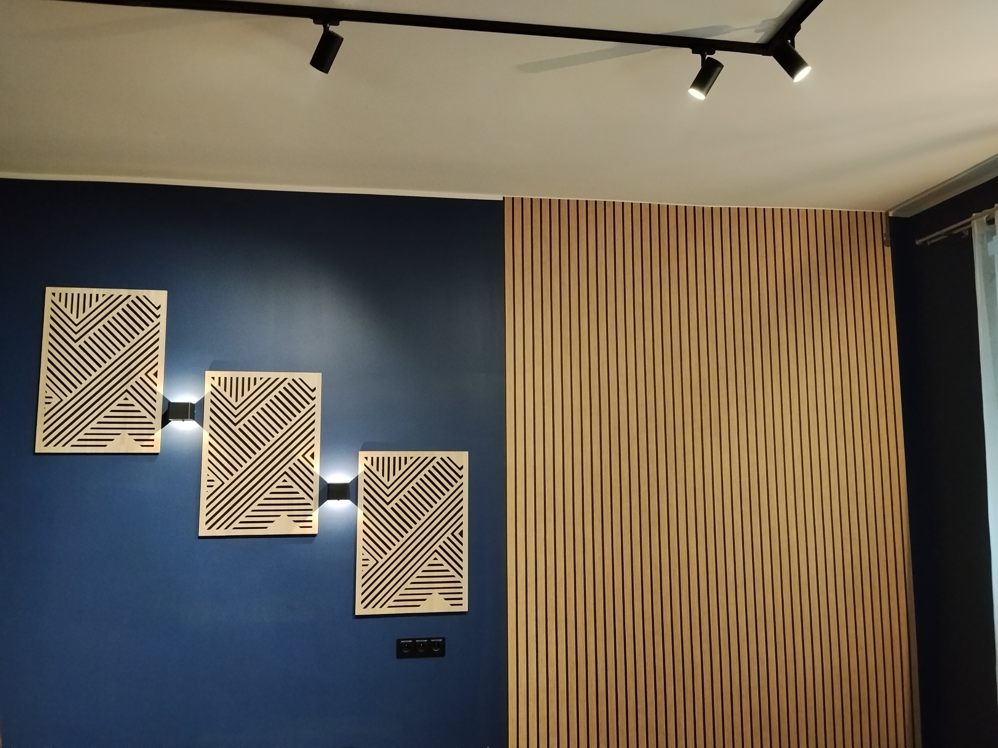 Niebieska ściana z trzema podświetlanymi panelami dekoracyjnymi i drewnianą ścianką lamelową. Oświetlenie sufitowe punktowe.