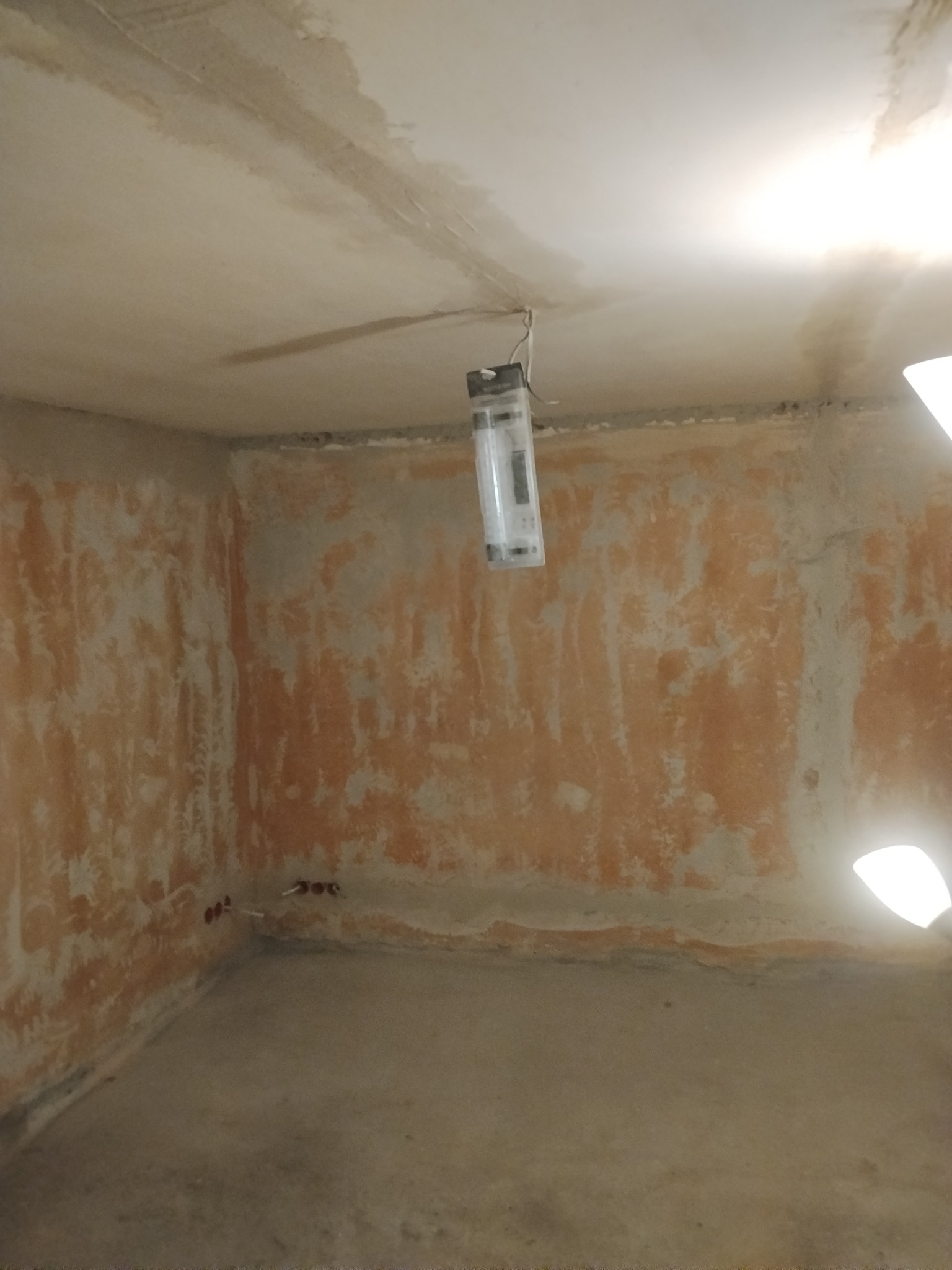 Pokój w trakcie remontu: ściany z resztkami farby, widoczne instalacje elektryczne, surowy sufit z wiszącą lampą. Przygotowanie do dalszych prac wykończeniowych.