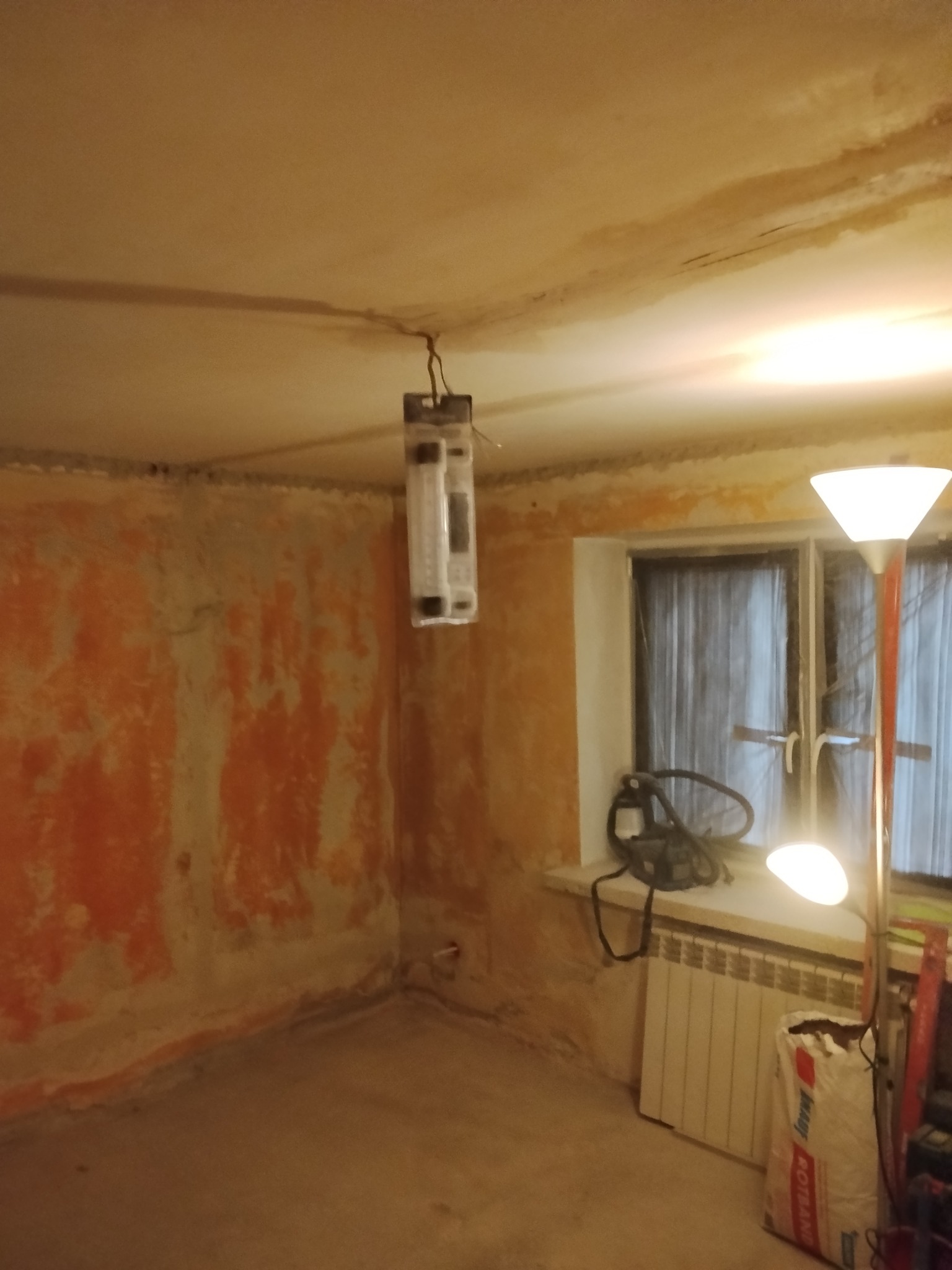 Pokój w trakcie remontu: ściany z resztkami farby, sufit z plamami, wisząca oprawa oświetleniowa w folii, grzejnik i worek z zaprawą. Widok na okno z zasłoną.