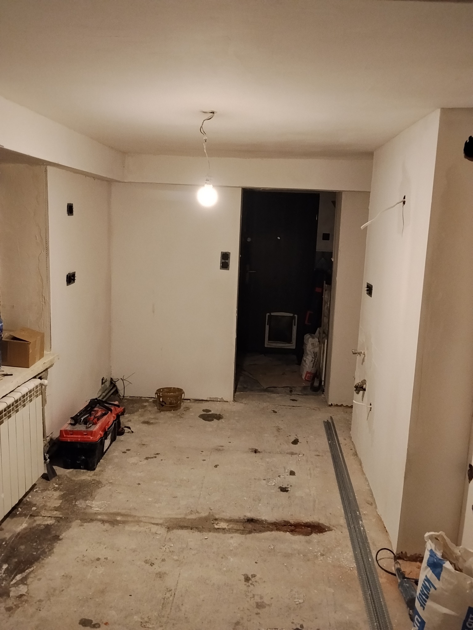 Wnętrze w trakcie remontu: surowe ściany, widoczne instalacje elektryczne, narzędzia i materiały budowlane na podłodze. Przygotowanie do wykończenia.