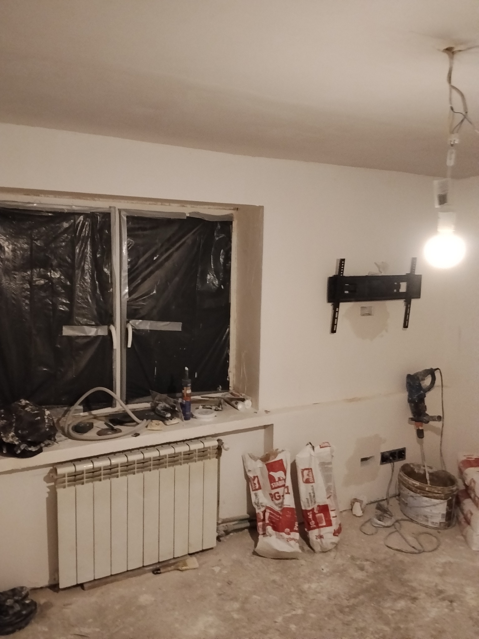 Pokój w trakcie prac wykończeniowych: okno zabezpieczone folią, zamontowany uchwyt TV, worki z zaprawą, wiadro z mieszadłem. Widoczne ślady po gładzi.