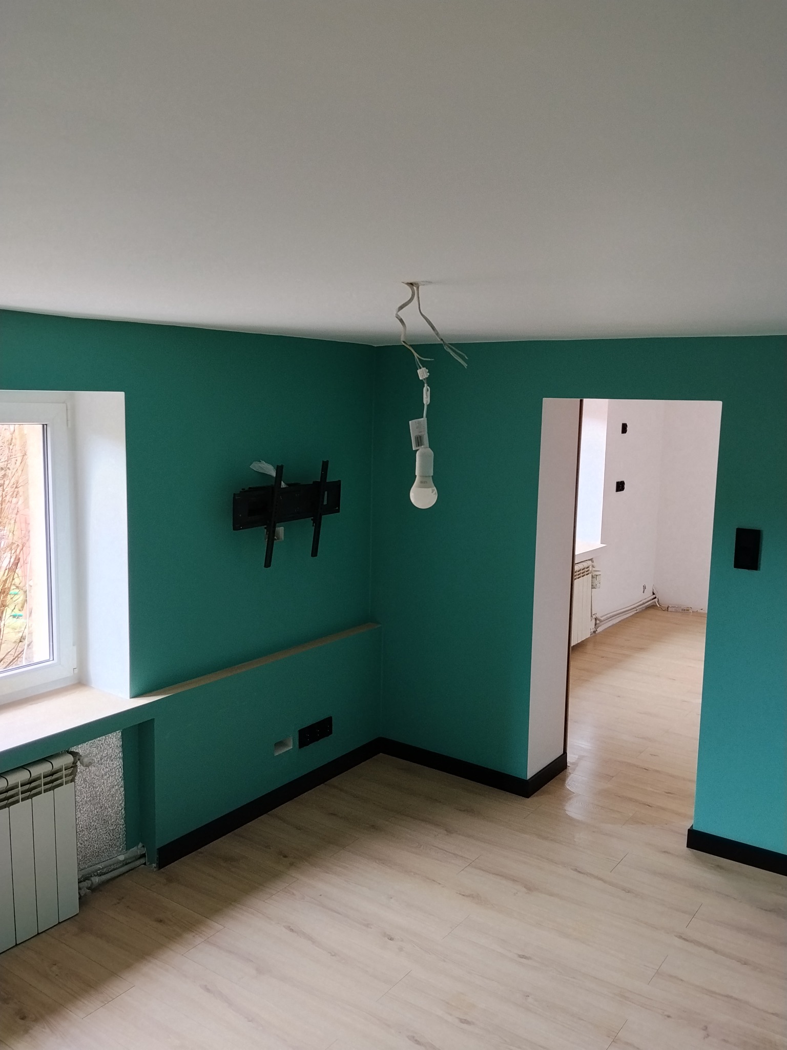 Pokój po remoncie: turkusowe ściany, jasna podłoga, białe okno i sufit. Widoczny grzejnik, uchwyt na TV i wisząca żarówka. W tle widok na kolejne, białe pomieszczenie.