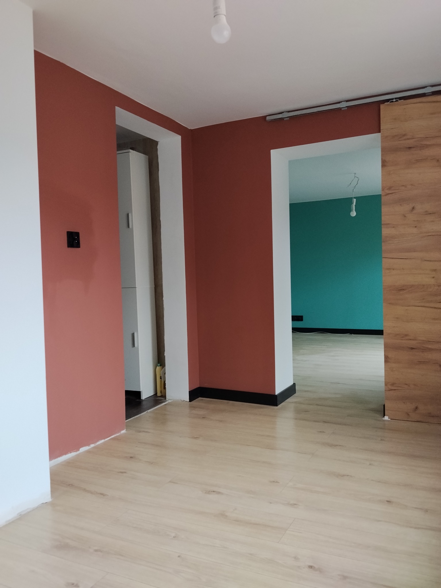 Wnętrze po malowaniu ścian na ceglasty kolor i położeniu jasnej podłogi. Widoczne drzwi przesuwne i fragment ściany w odcieniu turkusowym w kolejnym pomieszczeniu.