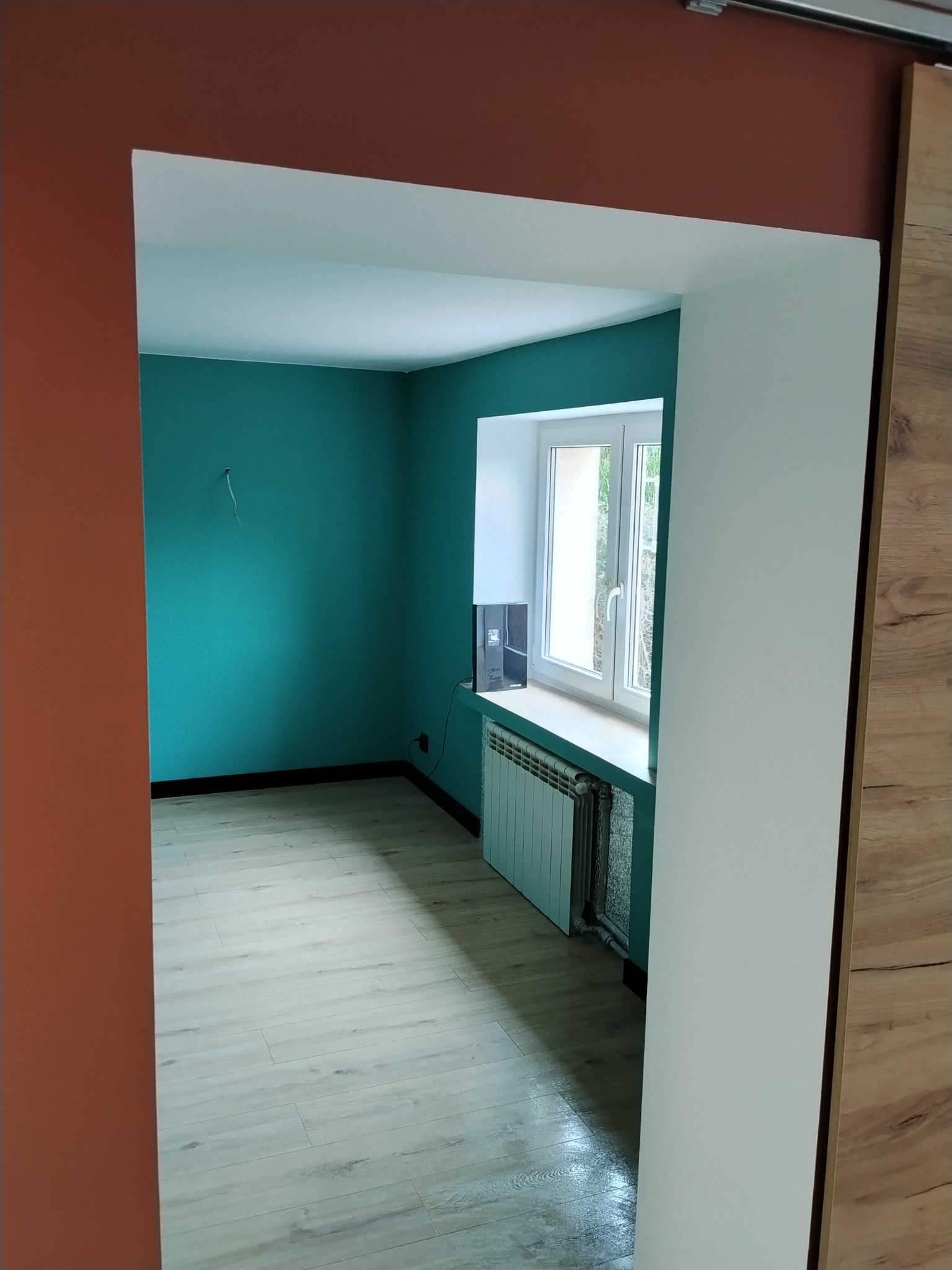 Pokój po remoncie: turkusowe ściany, jasna podłoga, białe okno z grzejnikiem. Widok z korytarza o pomarańczowej ścianie i drewnianych drzwiach przesuwnych.