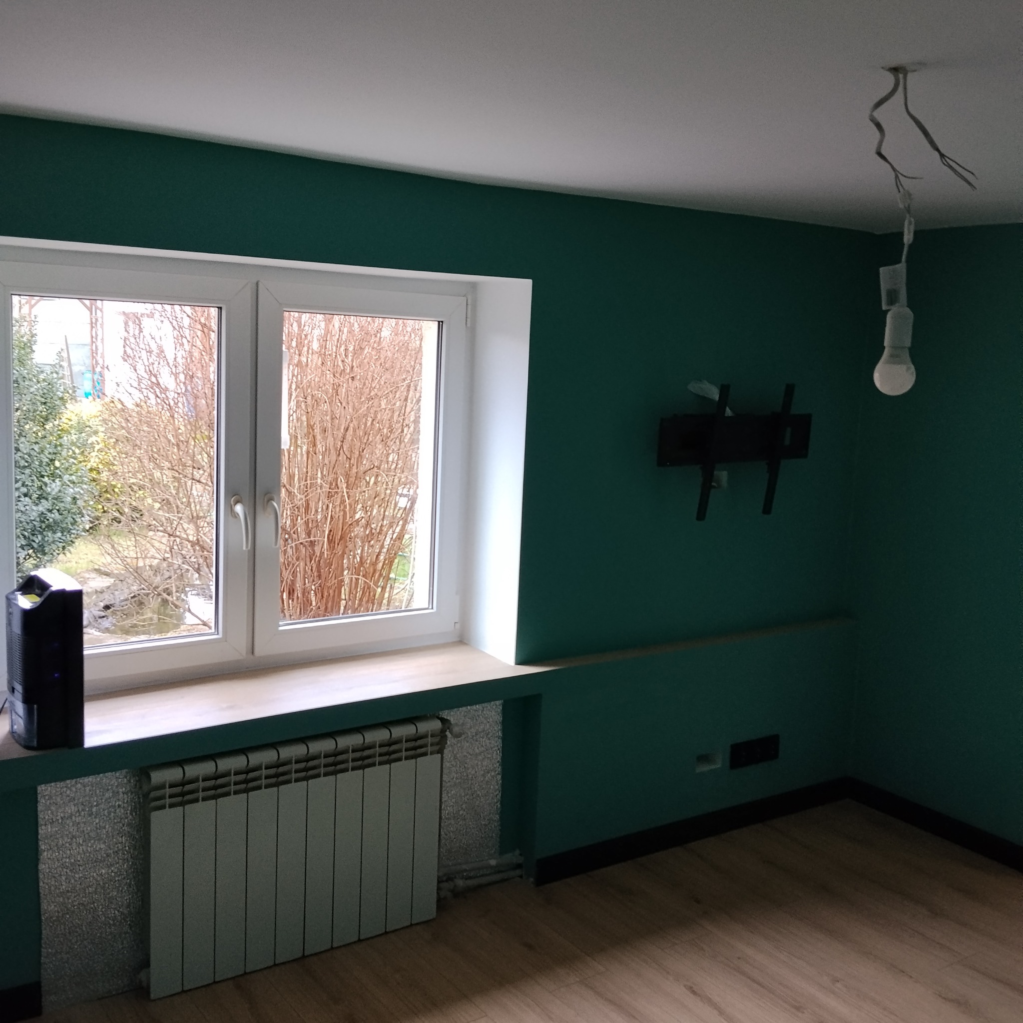 Pokój po malowaniu na butelkową zieleń w Łodzi. Widoczne okno, grzejnik, zamontowany uchwyt na telewizor i wisząca żarówka. Ściany pomalowane na jednolity kolor.