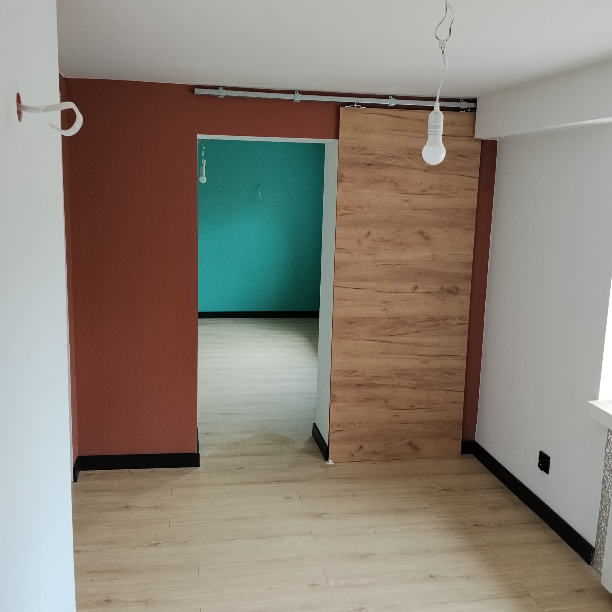 Pokój po remoncie: drewniane panele podłogowe, przesuwne drzwi z drewnianym wzorem, ściany w kolorach terakoty i turkusu. Widoczna surowa żarówka.