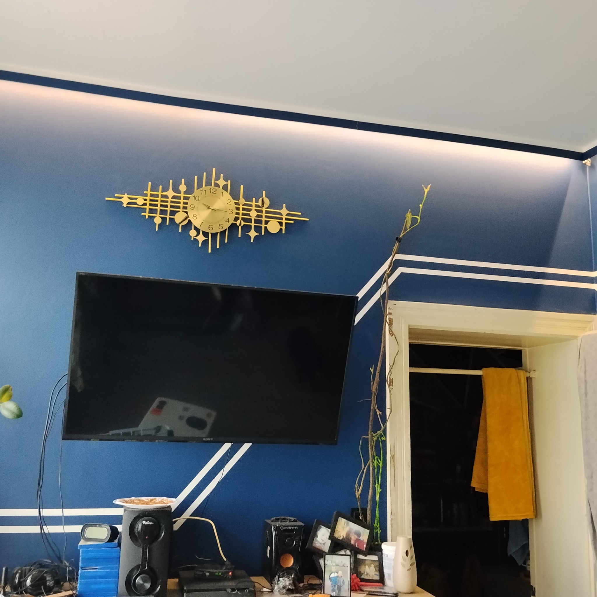 Niebieska ściana z dekoracyjnymi białymi pasami, złoty zegar w stylu retro, telewizor i rośliny. Nowoczesne wykończenie wnętrza z akcentami kolorystycznymi.