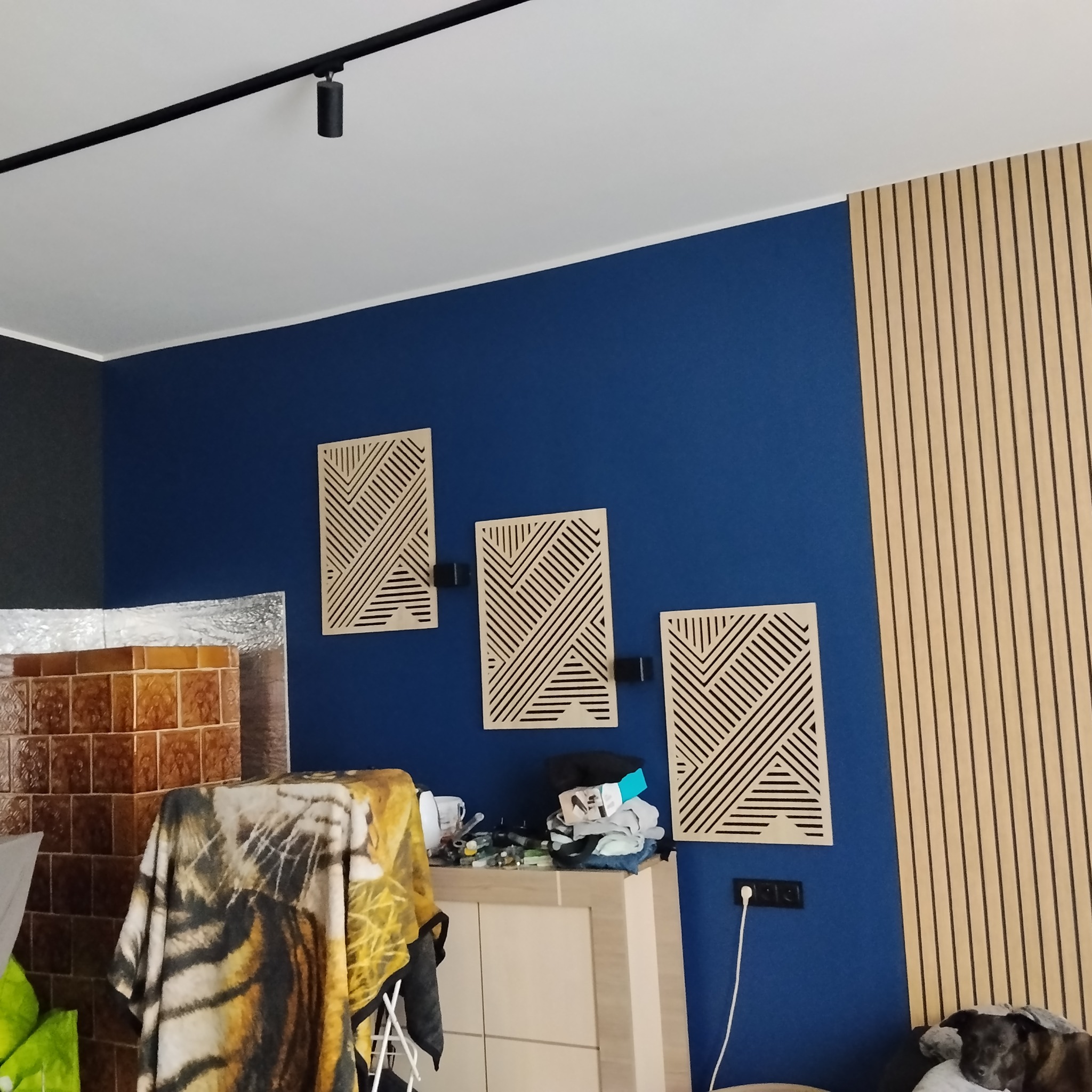 Niebieska ściana z trzema dekoracyjnymi panelami z geometrycznym wzorem, drewniana ściana z pionowymi listwami i fragment pokoju z widocznym grzejnikiem.