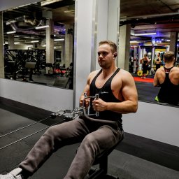 Damian Jankowski - Mężczyzna w czarnym tank topie GymBeam ćwiczy na maszynie do wiosłowania w siłowni, widoczny w lustrze drugi mężczyzna w tank topie. Buty Nike Air Jordan na nogach.