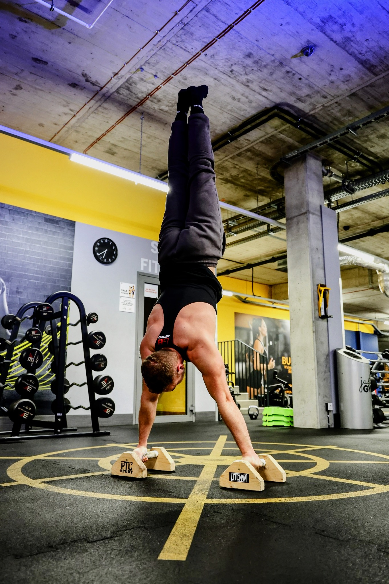 Mężczyzna w czarnej koszulce i dresach wykonuje stójkę na rękach na drewnianych platformach w siłowni z żółtymi akcentami i sprzętem do ćwiczeń.