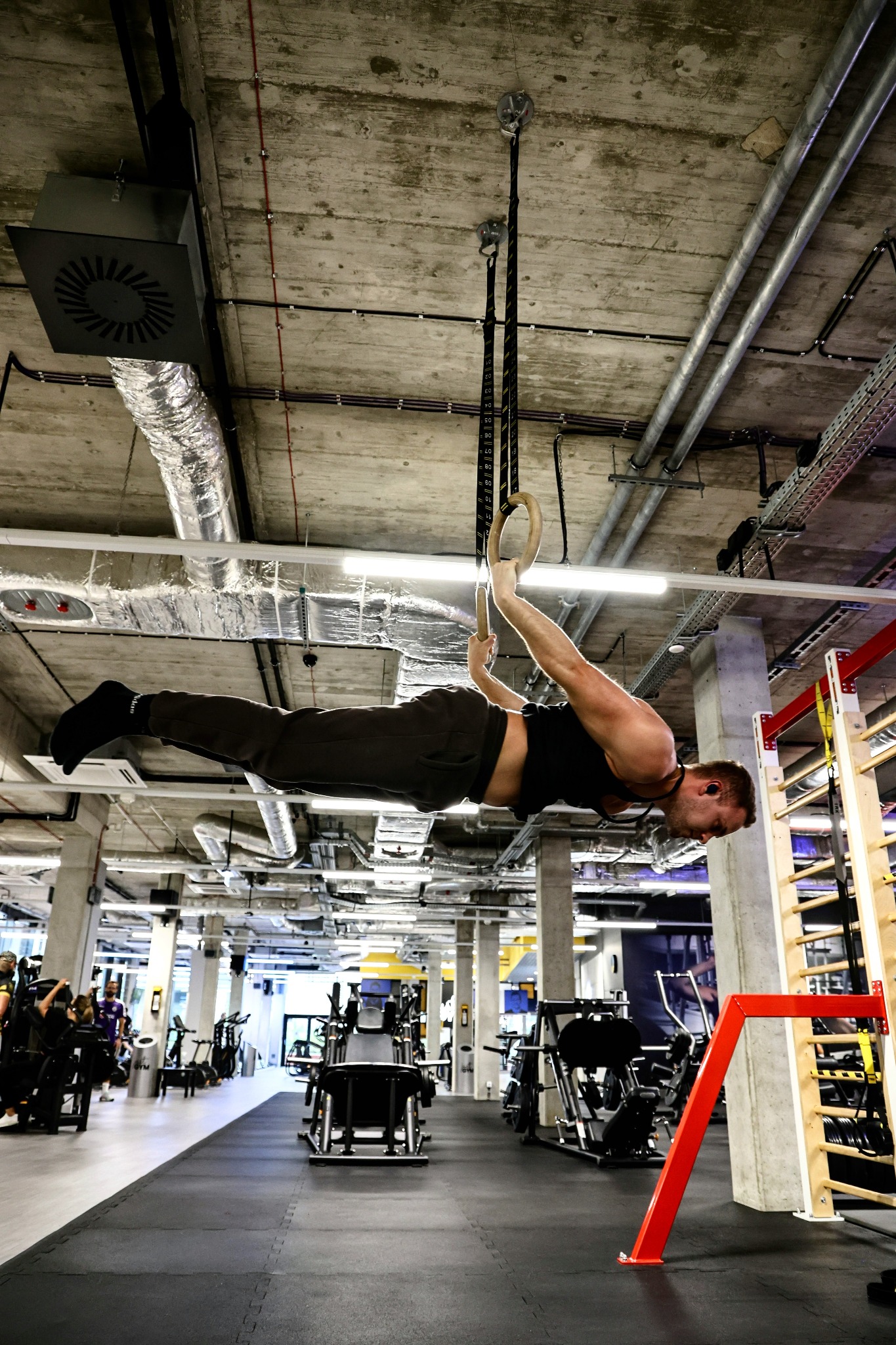 Mężczyzna w czarnym stroju wykonuje ćwiczenie na kółkach gimnastycznych w nowoczesnej siłowni z widocznymi instalacjami na suficie i sprzętem do ćwiczeń.