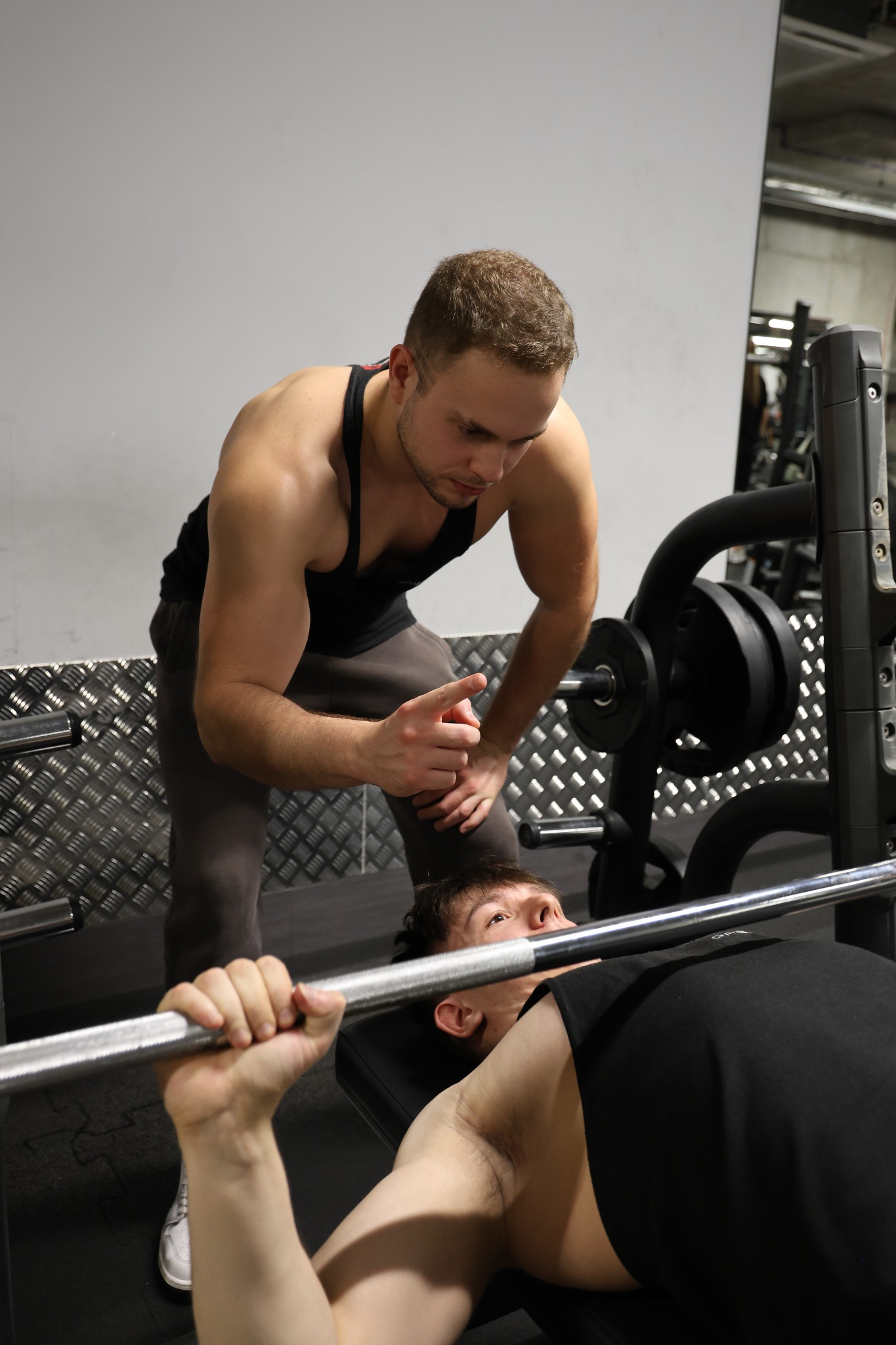 Mężczyzna ćwiczy wyciskanie sztangi na ławce pod okiem trenera, który udziela wskazówek w siłowni. Widoczne obciążenia i metalowa konstrukcja.