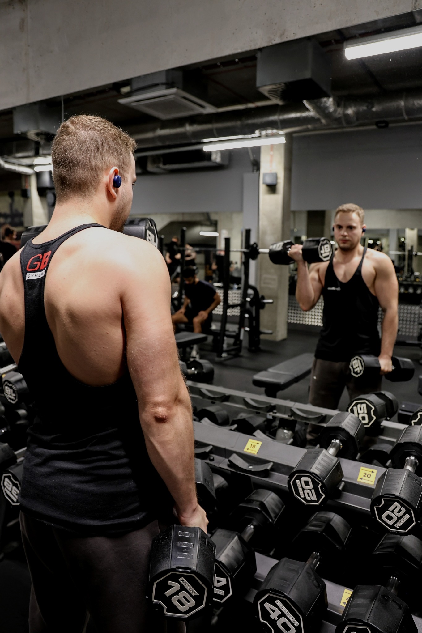 Mężczyzna z słuchawkami w czarnej koszulce na siłowni, trzymający hantle, widoczny w lustrze podczas ćwiczeń bicepsów.