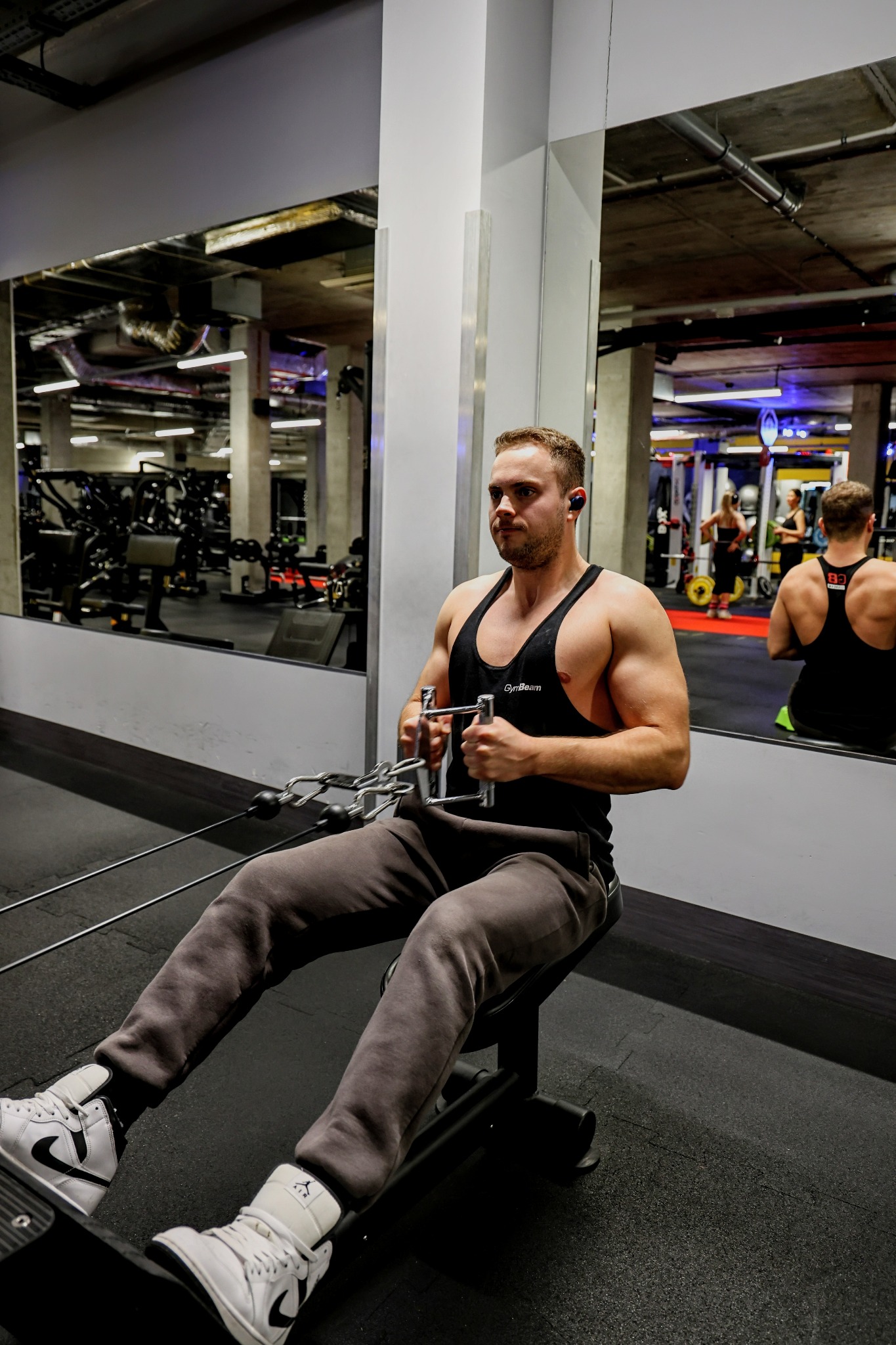 Mężczyzna w czarnym tank topie GymBeam ćwiczy na maszynie do wiosłowania w siłowni, widoczny w lustrze drugi mężczyzna w tank topie. Buty Nike Air Jordan na nogach.