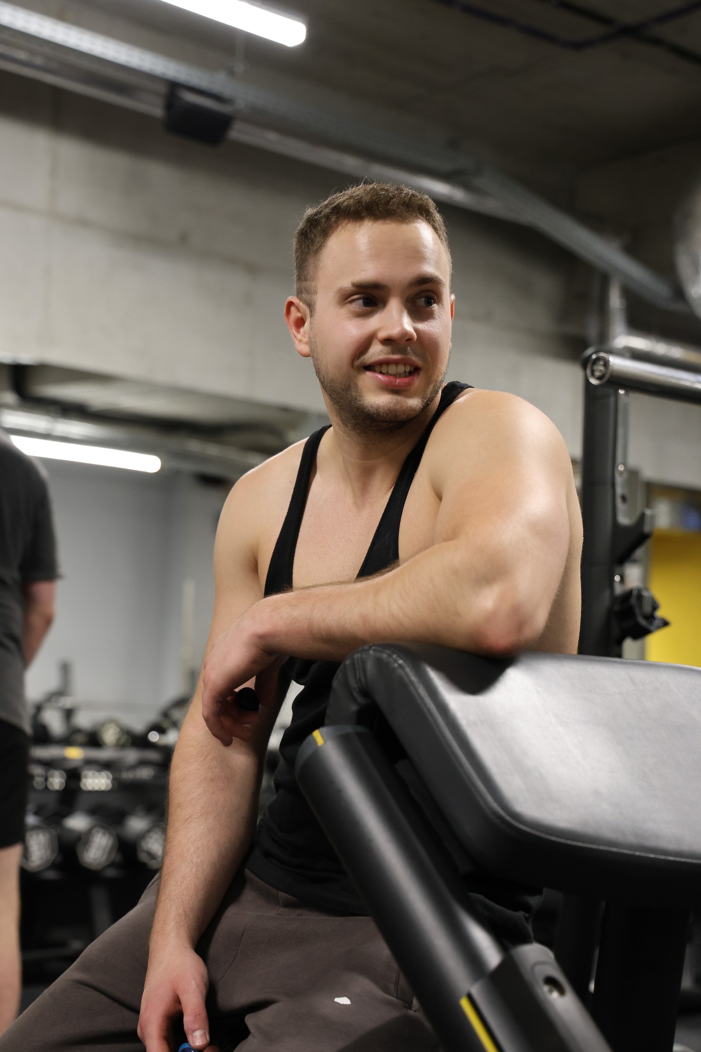 Mężczyzna w czarnej koszulce bez rękawów siedzi w siłowni, opierając się o sprzęt. W tle widać inne maszyny i osoby ćwiczące. Lekki uśmiech na twarzy.