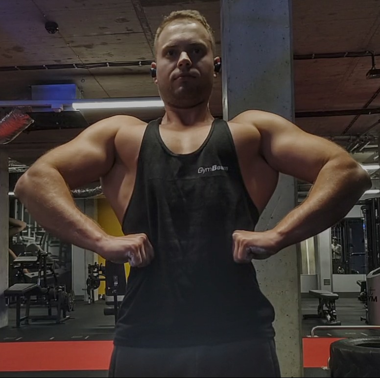 Mężczyzna w czarnej koszulce na siłowni prezentuje muskulaturę, z widocznym logo GymBeam na koszulce. W tle sprzęt do ćwiczeń i betonowy filar.