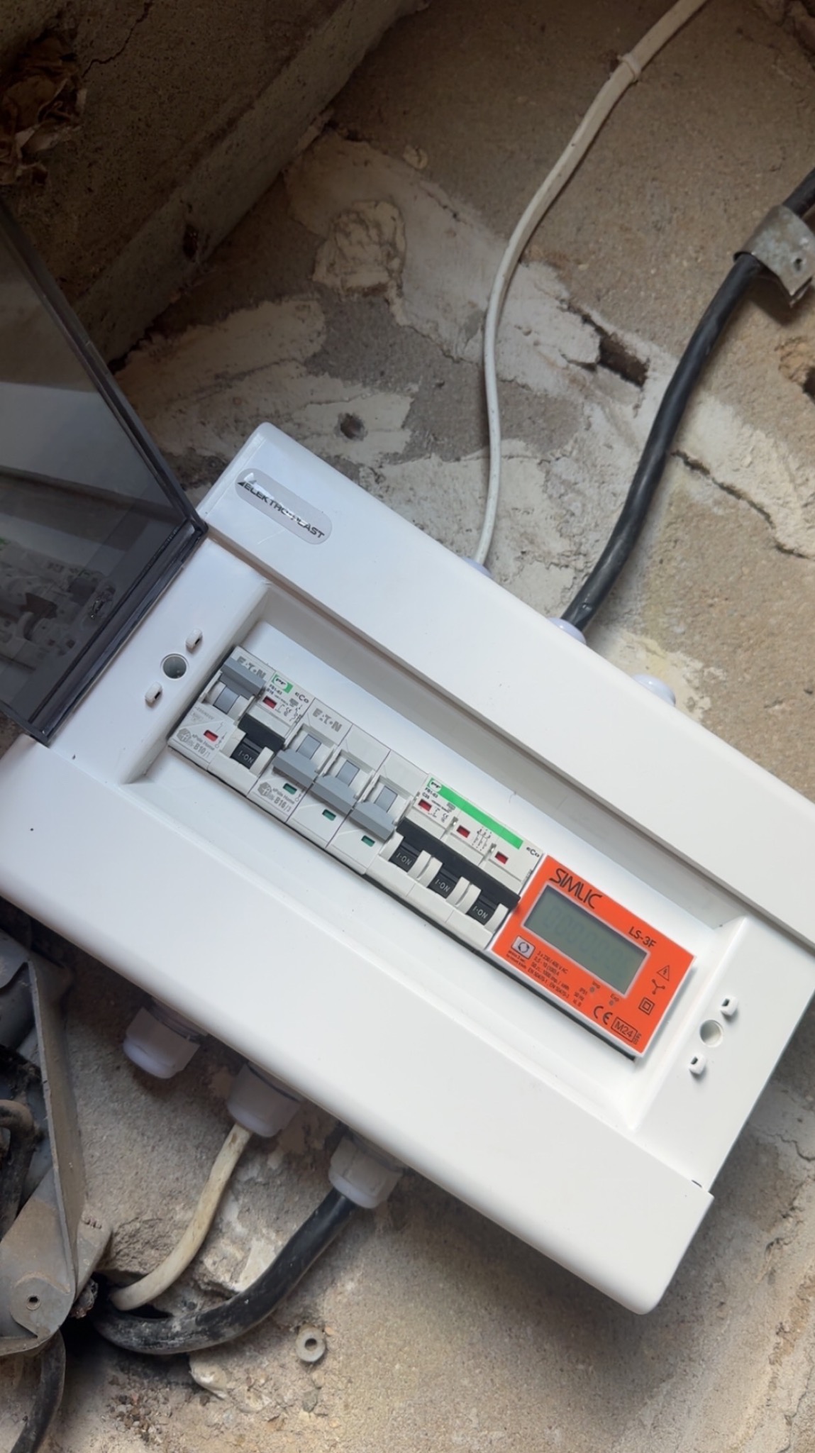 Otwarta skrzynka elektryczna z bezpiecznikami Eaton i miernikiem SIMLIC LS-3F, zamontowana na szarej, surowej ścianie. Widoczne przewody elektryczne.