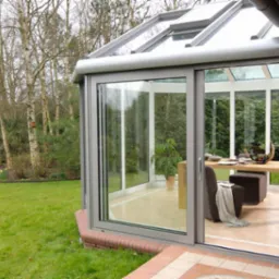 Szklana oranżeria z przesuwnymi drzwiami, widok z zewnątrz na drewniany stół z owocami i krzesło wewnątrz, ogród z drzewami w tle.