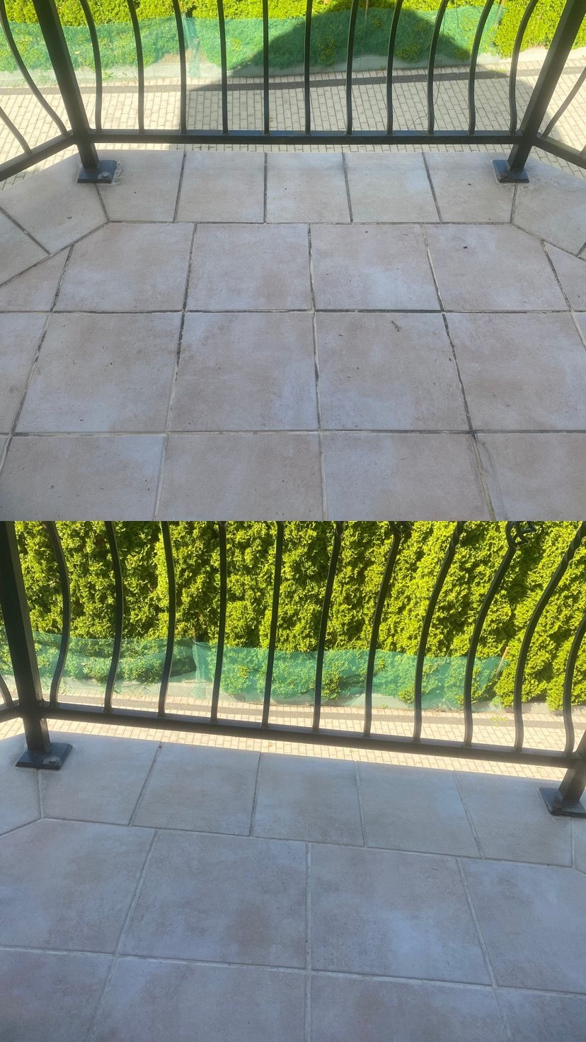 Balkon z jasnymi płytkami podłogowymi i czarną, ozdobną balustradą. W tle widoczna zielona roślinność. Podział kadru na dwie części.