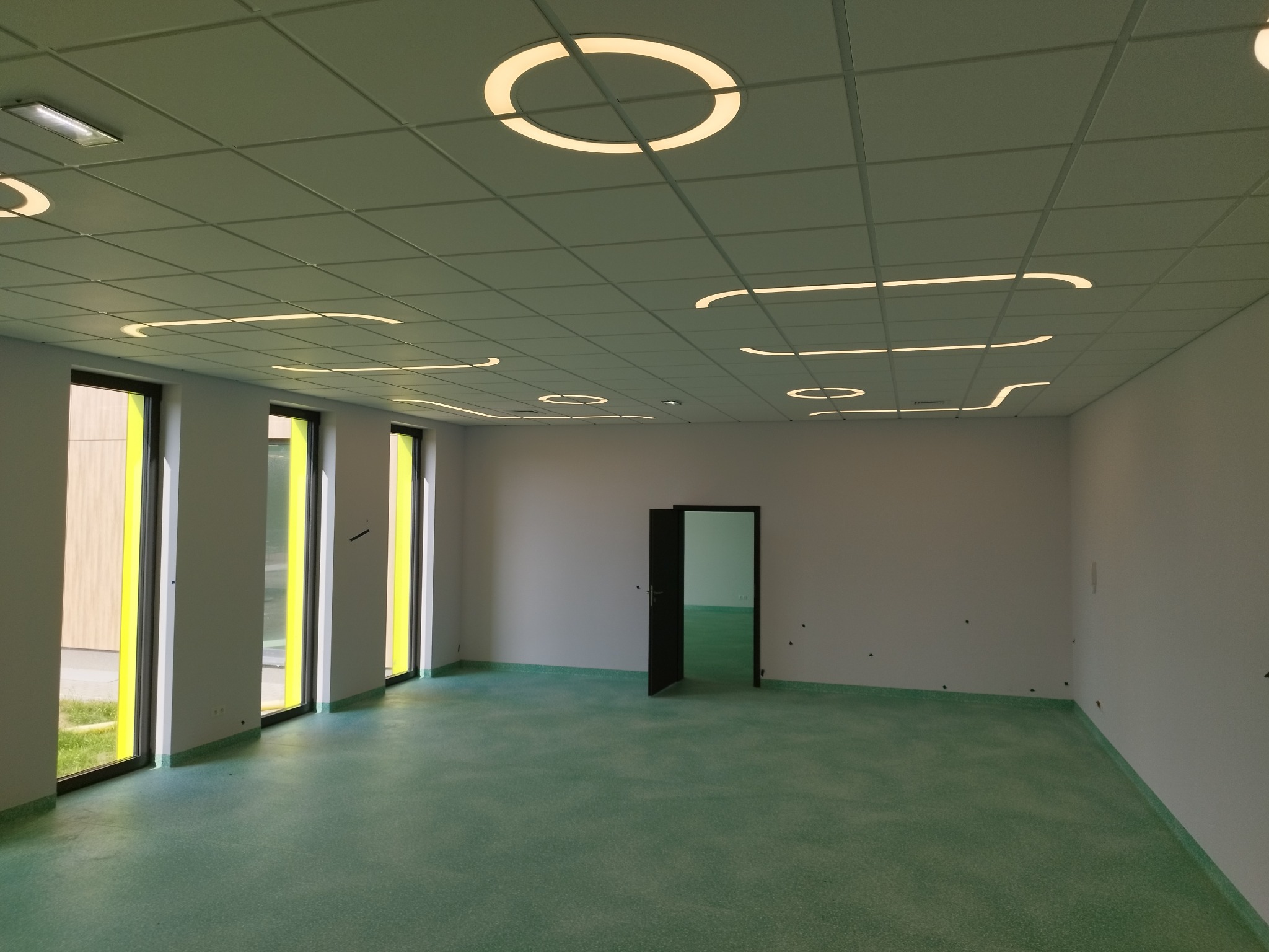 Jasne wnętrze biura z zieloną podłogą i designerskim oświetleniem sufitowym. Widoczne okna z żółtymi ramami i otwarte drzwi. Minimalistyczny styl.