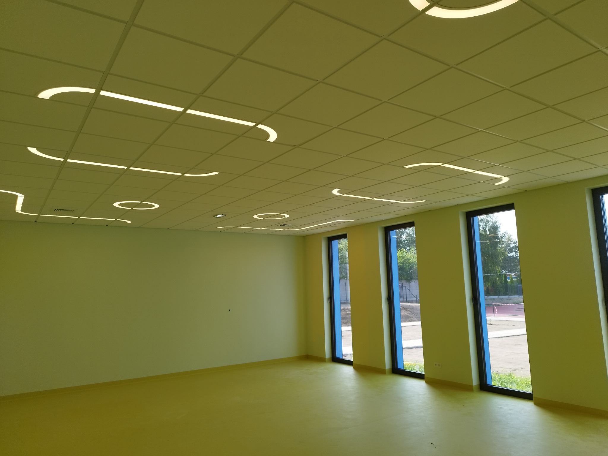 Pomalowane, puste pomieszczenie z podwieszanym sufitem i oryginalnym oświetleniem LED w kształcie linii i okręgów. Trzy okna z widokiem na zewnątrz.