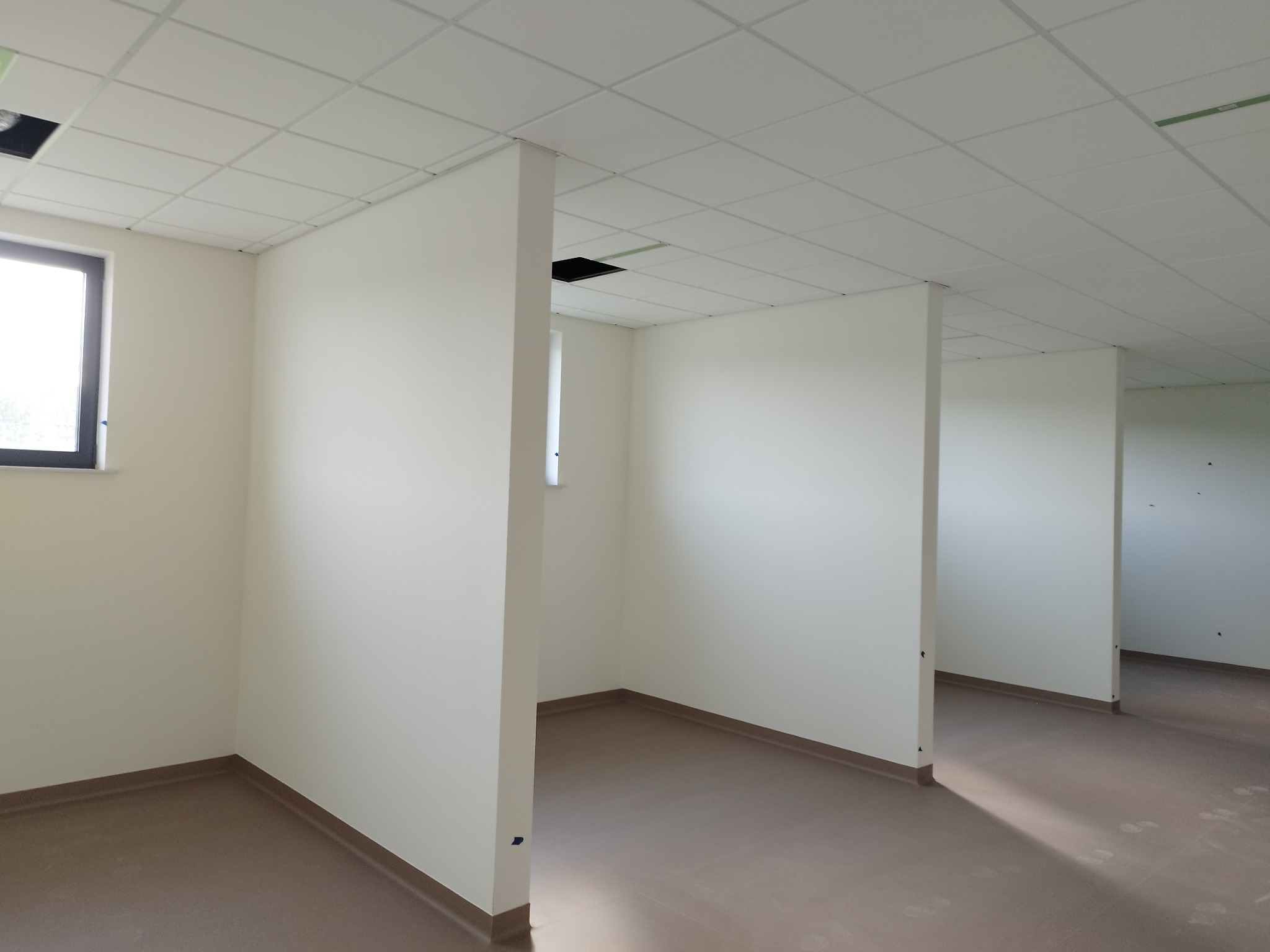 Jasne, świeżo pomalowane wnętrze biurowe z podwieszanym sufitem i panelami. Neutralna kolorystyka ścian i podłogi. Widoczne okno z czarną ramą.