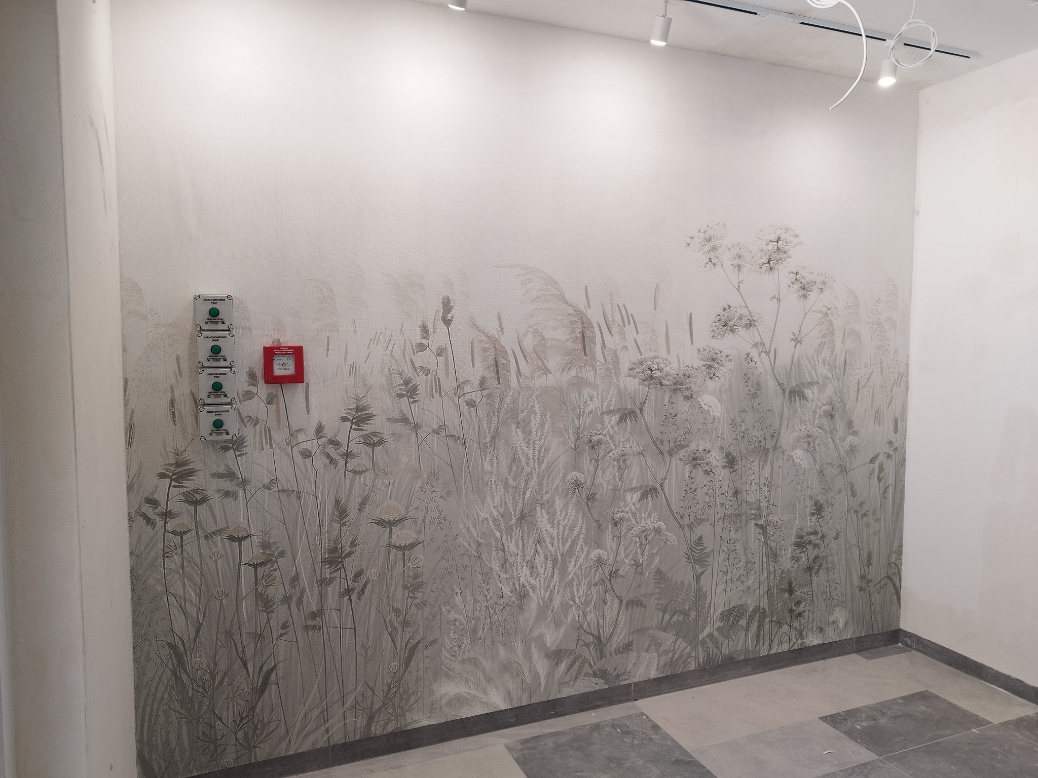 Ściana z subtelnym motywem łąki w stonowanych szarościach, z elementami instalacji elektrycznej po lewej stronie. Nowoczesne oświetlenie sufitowe.