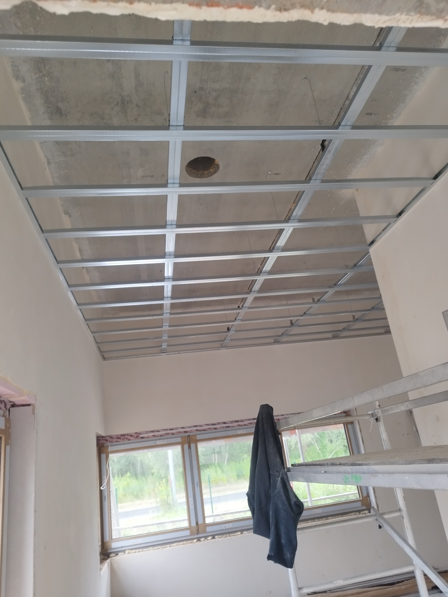 Metalowy stelaż sufitowy z widocznym otworem na instalację, zamontowany w surowym wnętrzu z oknami i rusztowaniem, przygotowany pod montaż płyt.