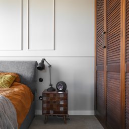 Sypialnia w Sopocie: łóżko z szarą ramą, drewniana szafka nocna z lampką, brązowa szafa z żaluzjowymi drzwiami. Ściana z białymi panelami dekoracyjnymi.