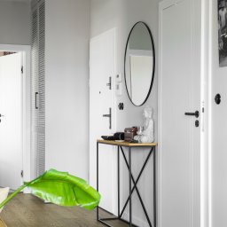 Jasne wnętrze przedpokoju z drewnianą podłogą, białymi drzwiami i minimalistyczną konsolą z dekoracjami. Oświetlenie szynowe i lustro dopełniają aranżację.