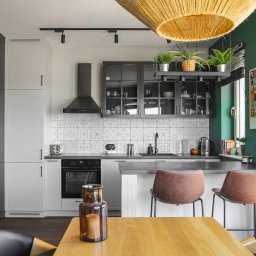 Nowoczesna kuchnia z geometrycznymi płytkami, czarnymi szafkami i drewnianym stołem. W tle zielona ściana i okno. Nad blatem wiszą rośliny doniczkowe.