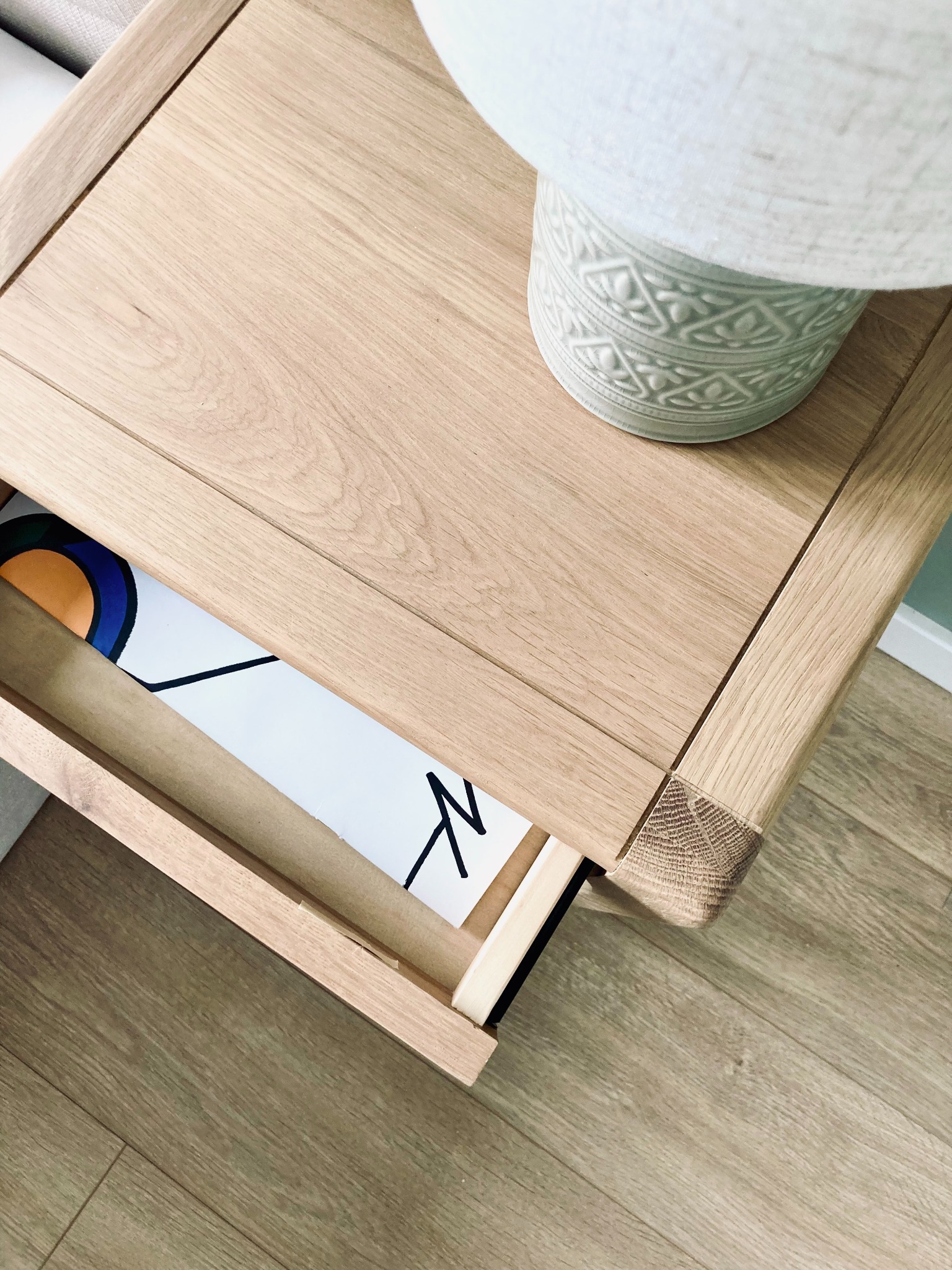 Drewniana szafka nocna z wysuniętą szufladą, na której stoi lampka nocna. W szufladzie widoczny plakat z abstrakcyjnym wzorem. Ujęcie z góry, naturalne światło.