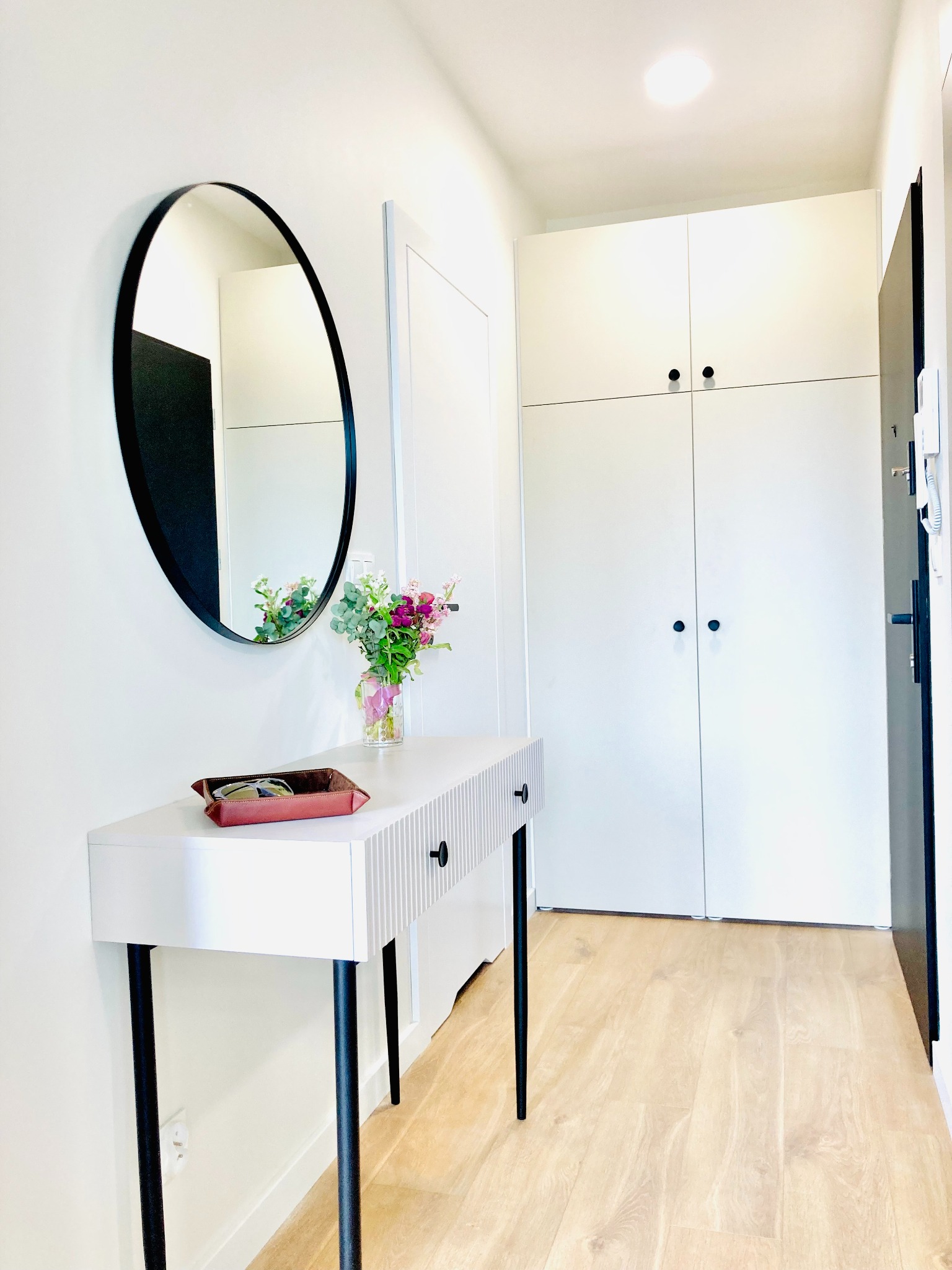 Nowoczesny przedpokój z białymi szafami, konsolą z kwiatami i okrągłym lustrem w czarnej ramie. Jasne panele podłogowe. Minimalistyczny design.