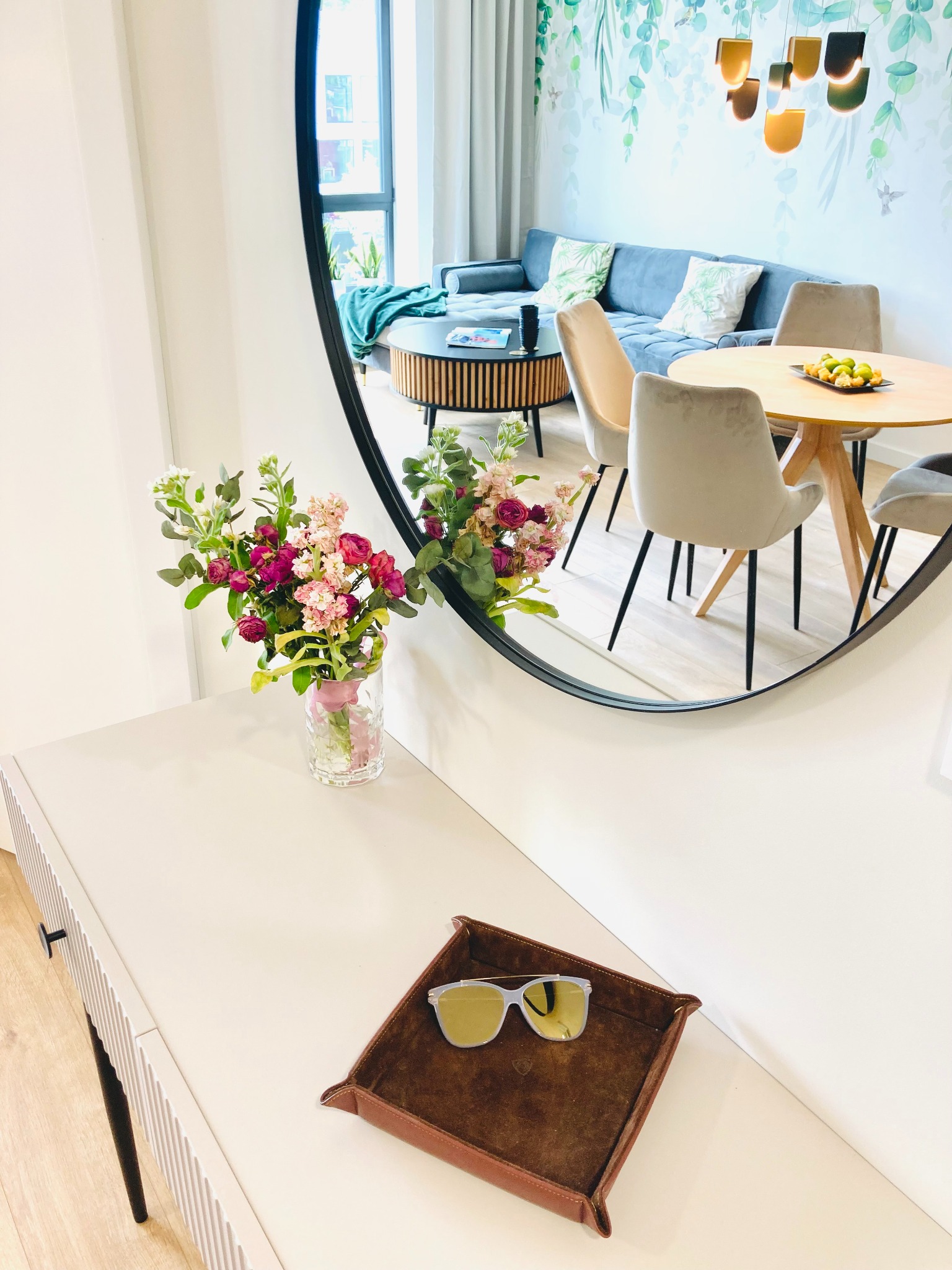 Nowoczesne wnętrze mieszkania odbite w okrągłym lustrze: salon z szarą sofą, drewnianym stołem i kwiatami na komodzie. Okulary leżą na tacy.