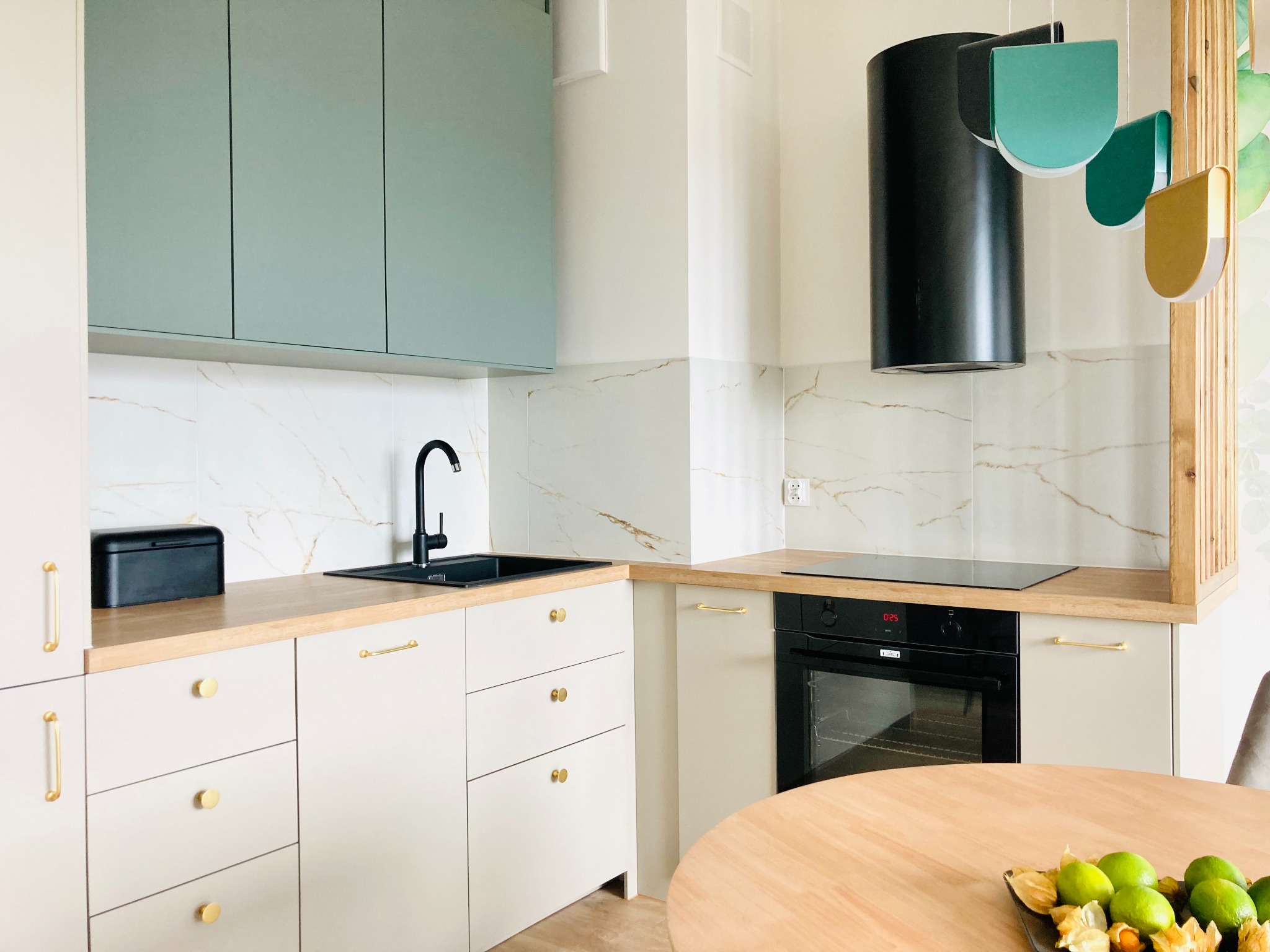 Jasna kuchnia z miętową zabudową górną i złotymi uchwytami. Na blacie czarny zlew i płyta indukcyjna. Okrągły stół z owocami na pierwszym planie.