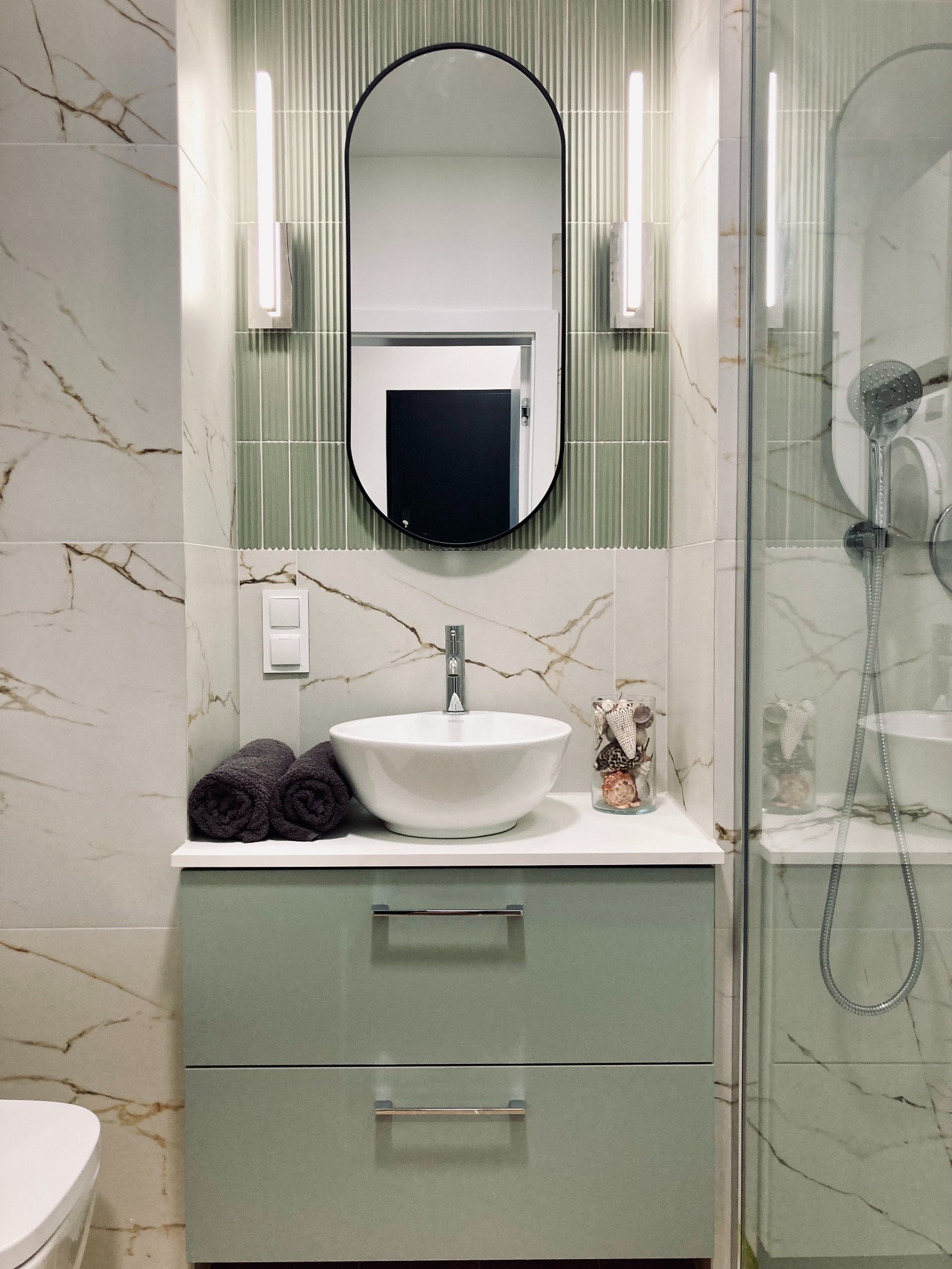 Nowoczesna łazienka z umywalką nablatową, szafką w odcieniach zieleni i lustrem w czarnej ramie. Marmurowe płytki i szklana kabina prysznicowa.