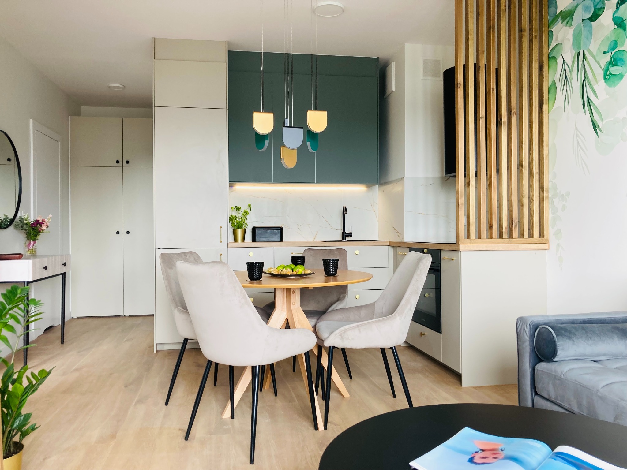 Jasne wnętrze z okrągłym stołem i krzesłami, zielona zabudowa kuchenna z marmurowym blatem, drewniana ścianka działowa i roślinny motyw na ścianie.