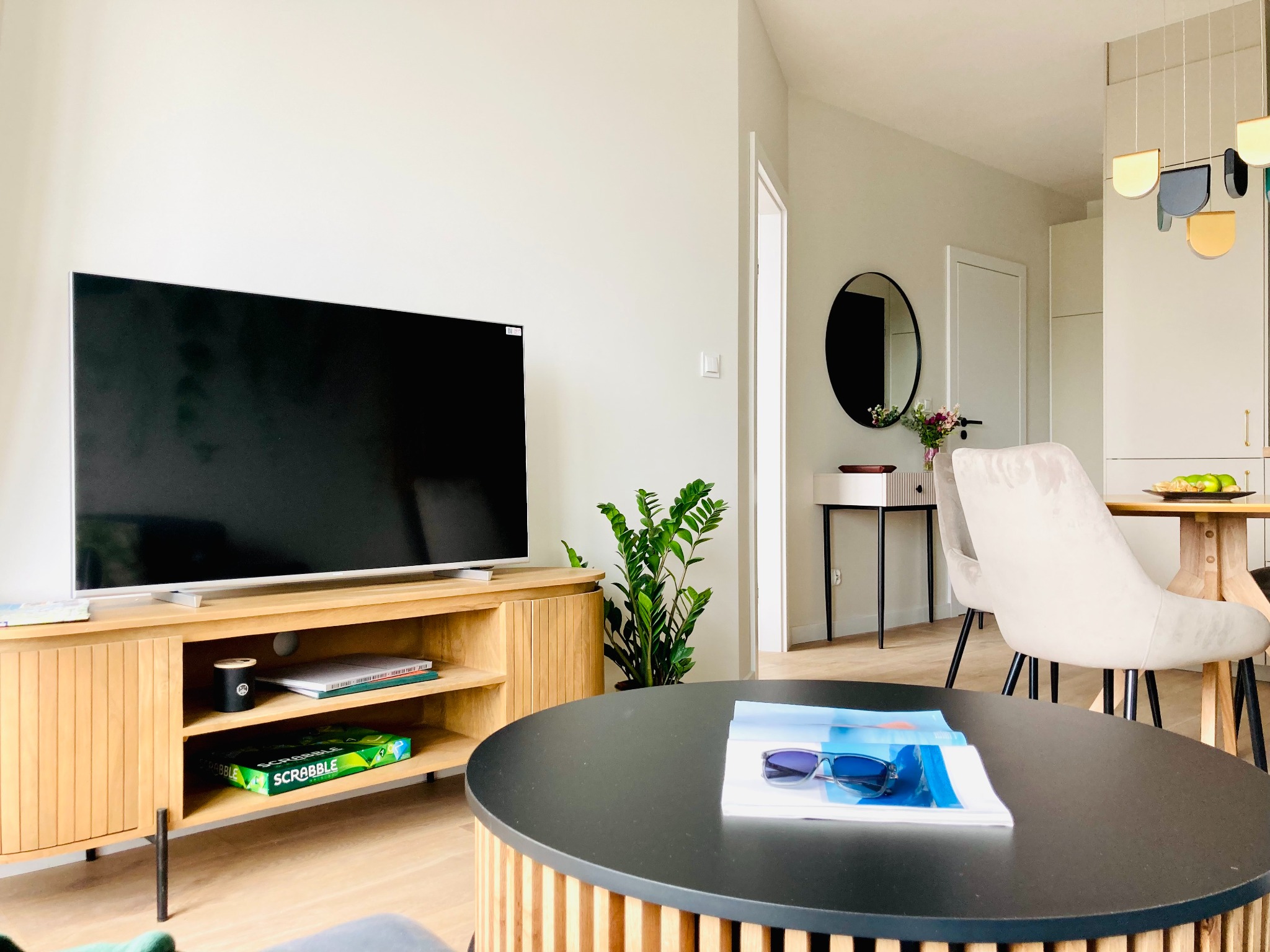 Jasne wnętrze salonu z drewnianą szafką RTV, okrągłym stolikiem kawowym i rośliną doniczkową. Minimalistyczny design, okrągłe lustro i designerska lampa.