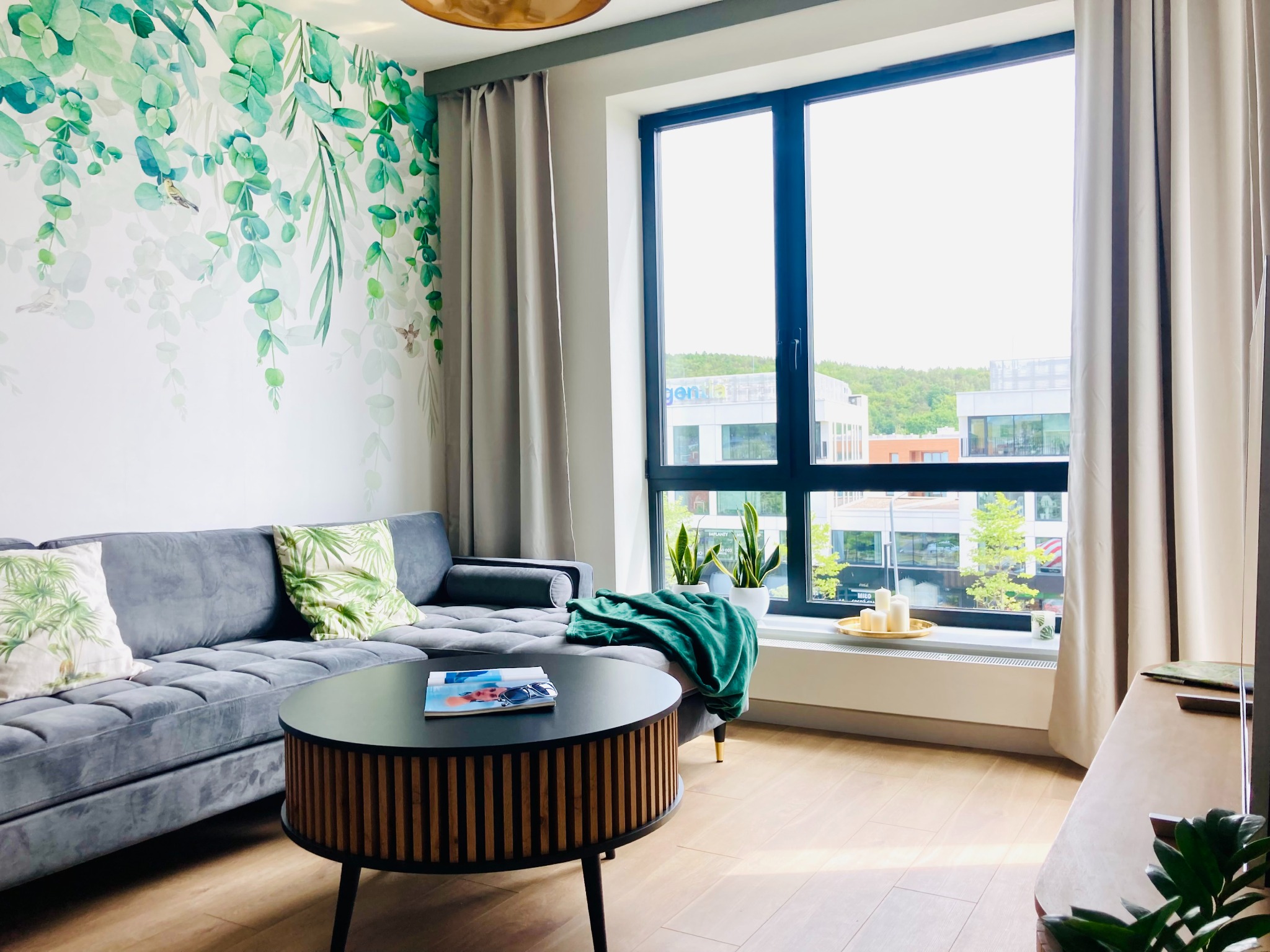 Stylowy salon z szarą sofą, okrągłym stolikiem kawowym i ścianą z motywem roślinnym. Duże okno z widokiem na budynki. Jasne drewniane podłogi.