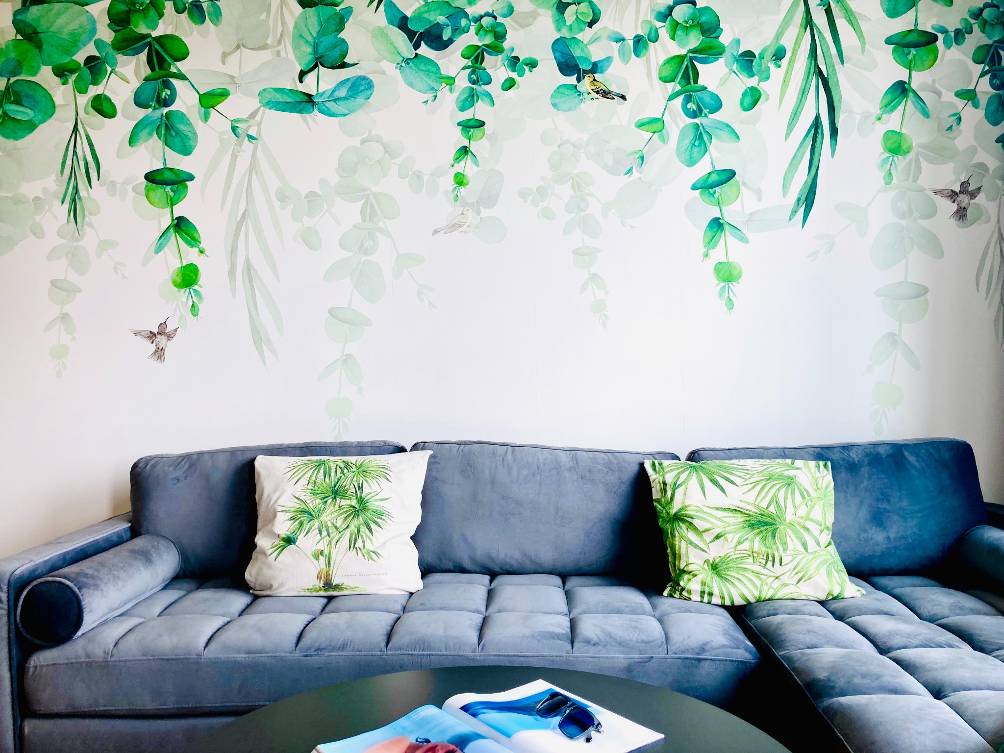 Szary narożnik z poduszkami w tropikalne wzory na tle ściany z motywem roślinnym i kolibrami. Nowoczesny design wnętrza z akcentem natury.