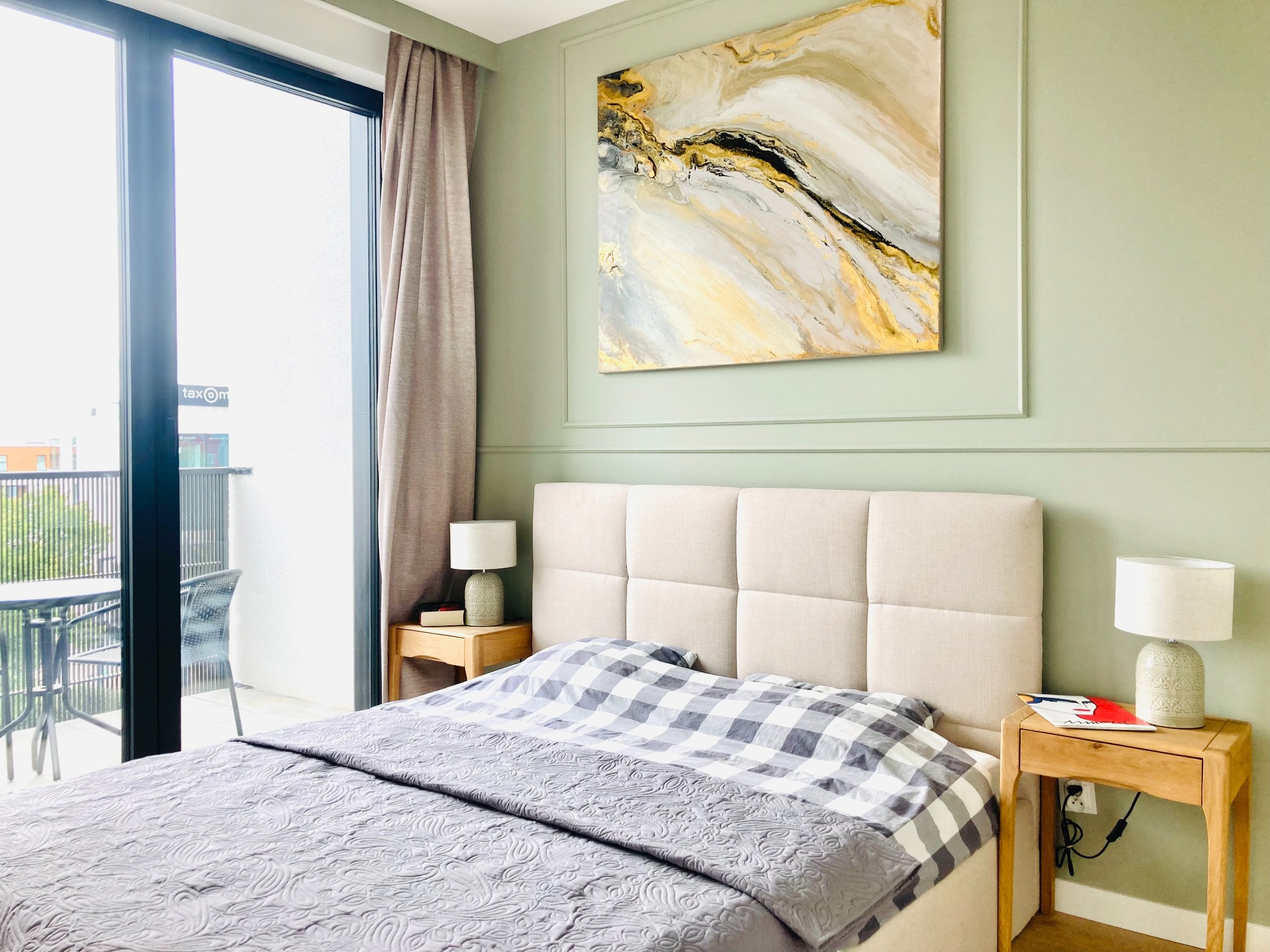 Stylowa sypialnia w Sopocie z łóżkiem, stolikami nocnymi i abstrakcyjnym obrazem na ścianie. Widok na balkon przez duże okno. Pastelowe kolory i nowoczesny design.