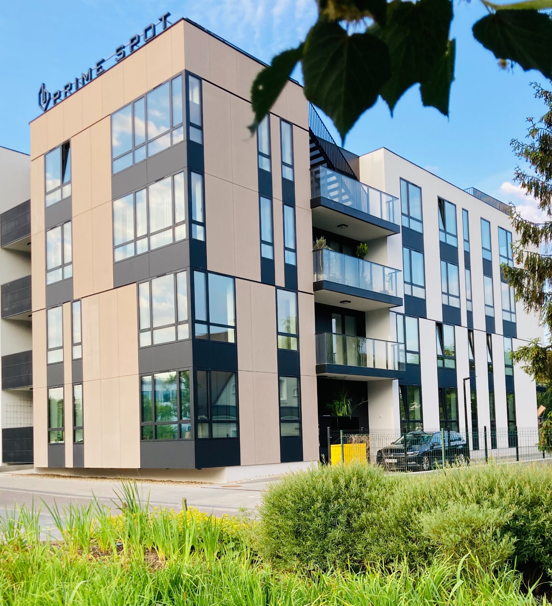 Nowoczesny budynek apartamentowy z balkonami i dużymi oknami, w odcieniach beżu, bieli i czerni, z zielenią w tle i logo 'Prime Spot' na szczycie.