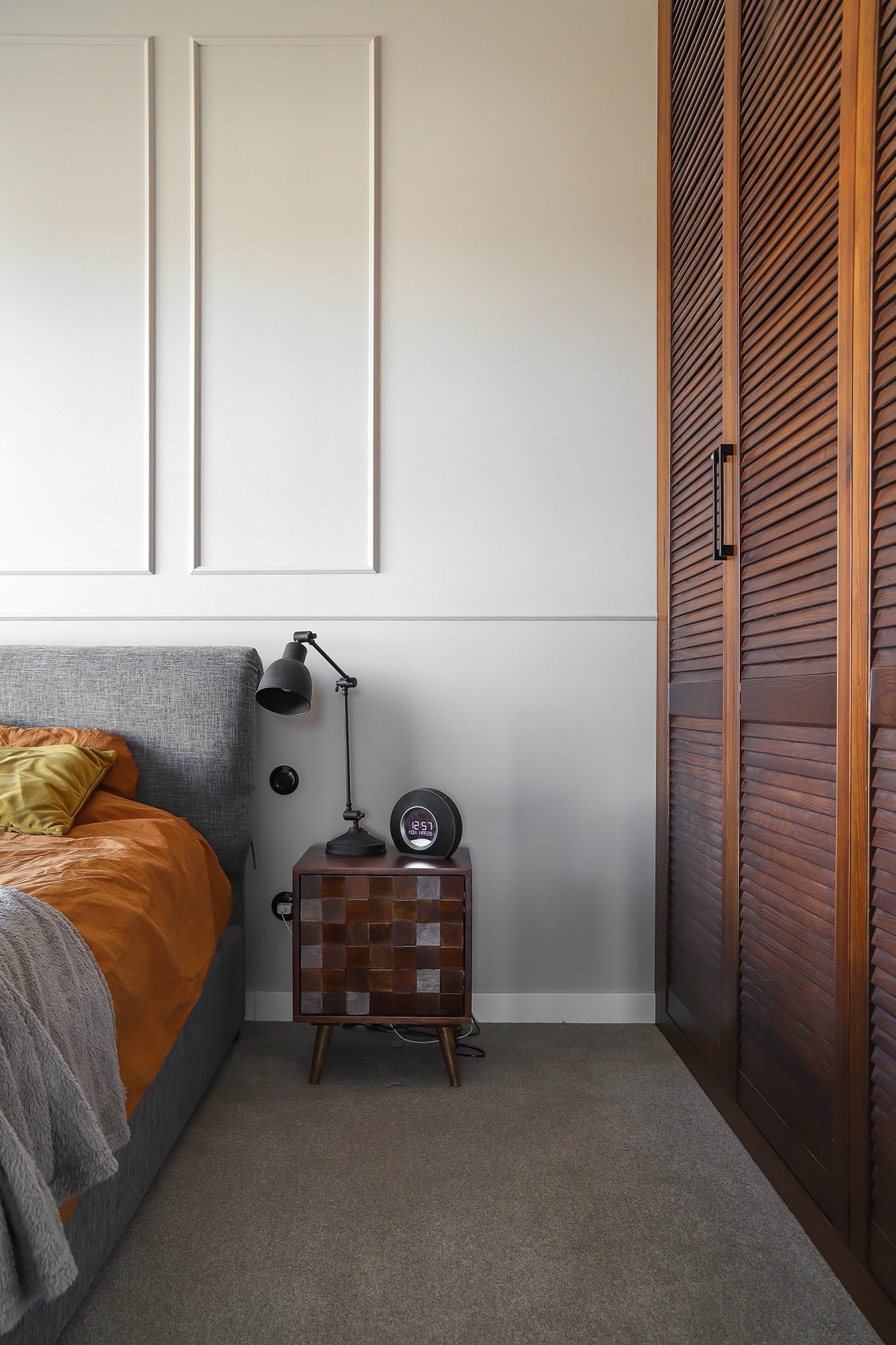 Sypialnia w Sopocie: łóżko z szarą ramą, drewniana szafka nocna z lampką, brązowa szafa z żaluzjowymi drzwiami. Ściana z białymi panelami dekoracyjnymi.