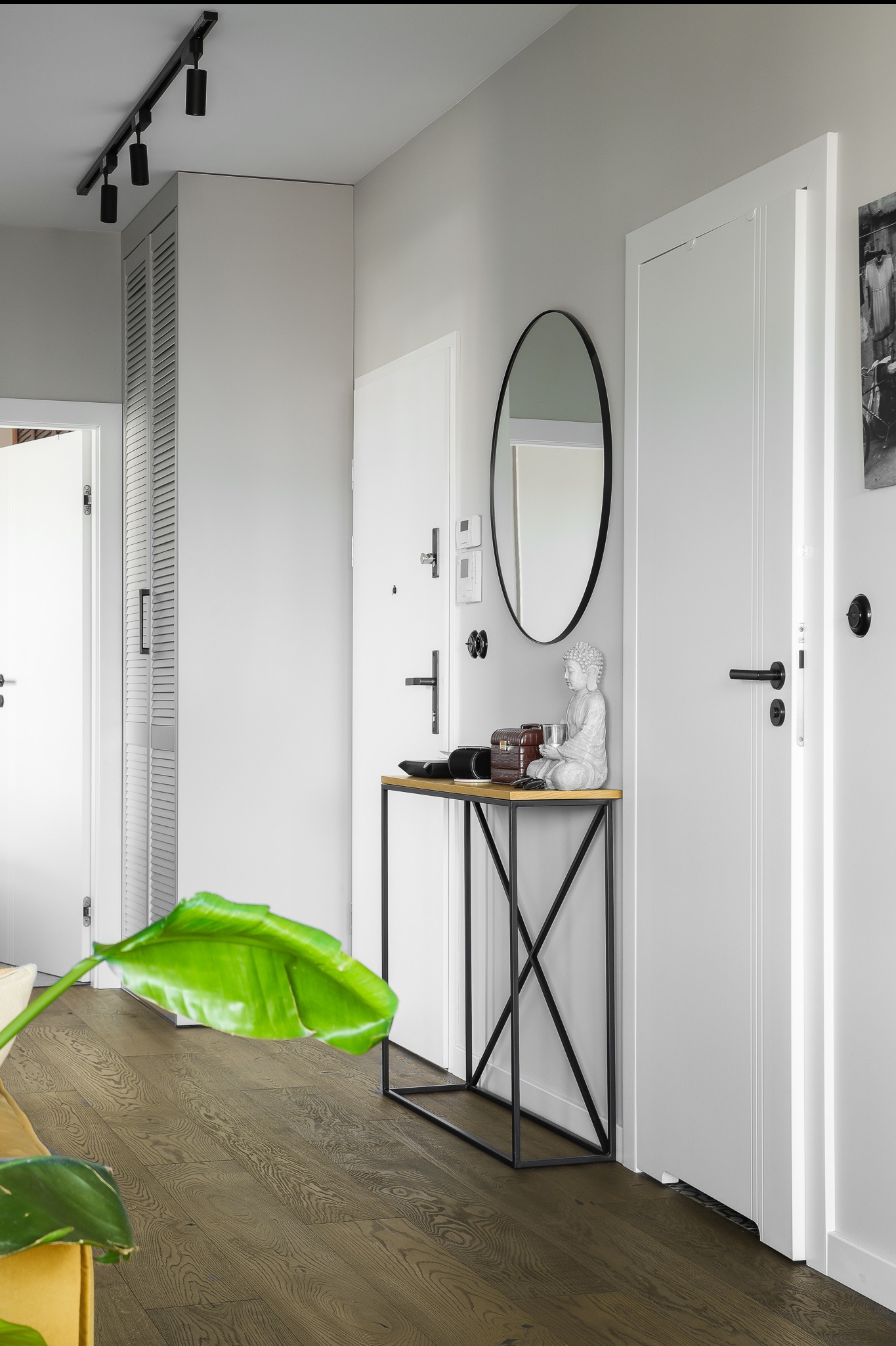 Jasne wnętrze przedpokoju z drewnianą podłogą, białymi drzwiami i minimalistyczną konsolą z dekoracjami. Oświetlenie szynowe i lustro dopełniają aranżację.