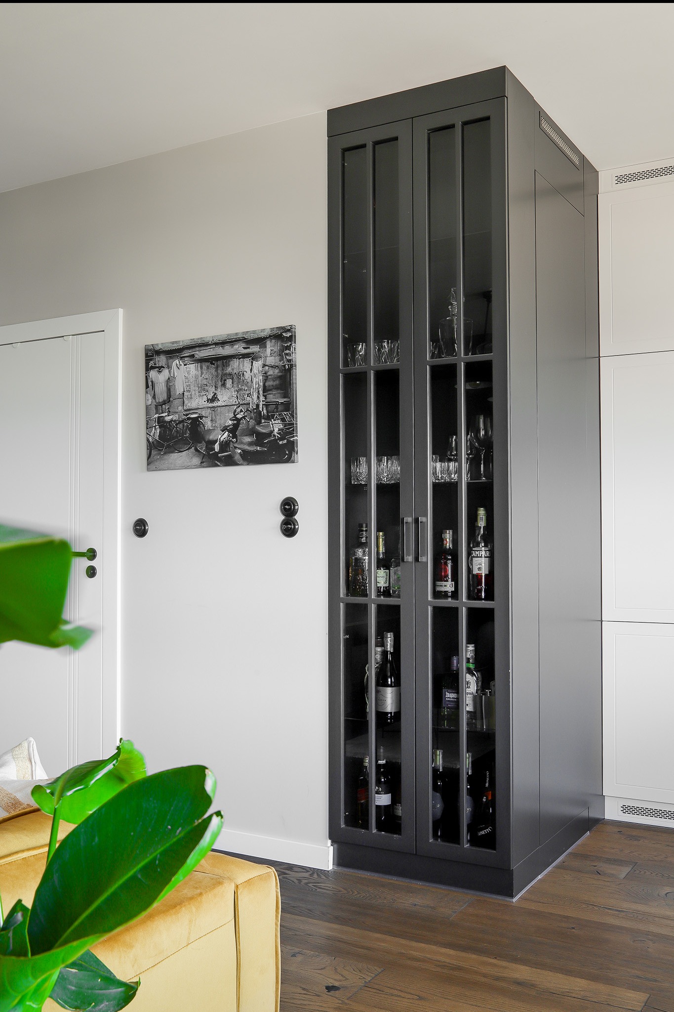 Szara, wysoka witryna z przeszkleniami, wypełniona szkłem i butelkami, wkomponowana w nowoczesne wnętrze z drewnianą podłogą i białymi ścianami.