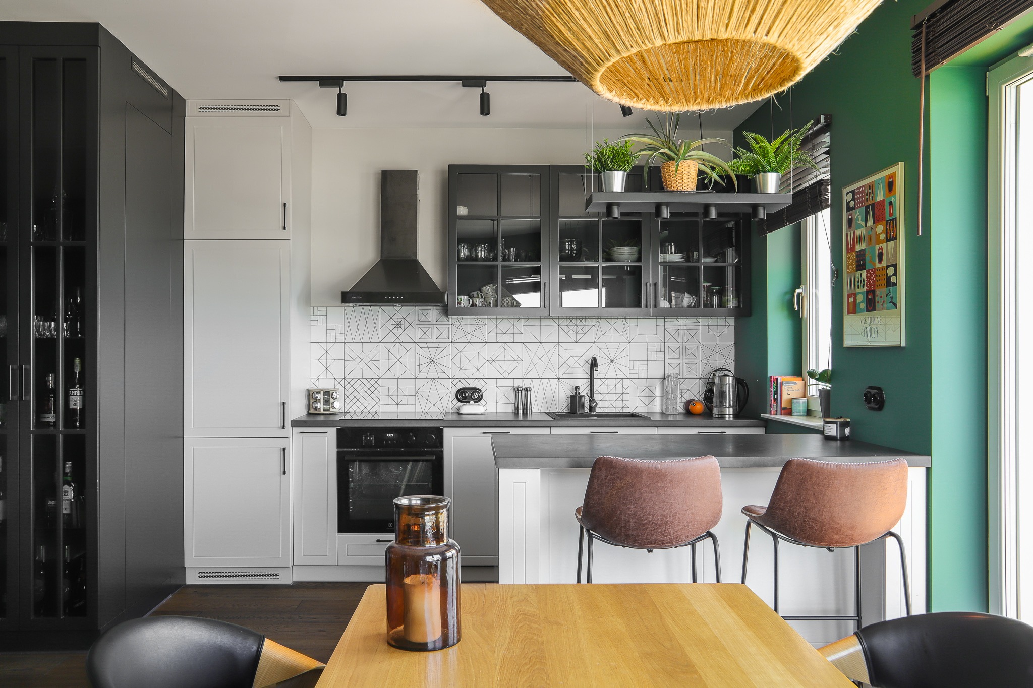 Nowoczesna kuchnia z geometrycznymi płytkami, czarnymi szafkami i drewnianym stołem. W tle zielona ściana i okno. Nad blatem wiszą rośliny doniczkowe.