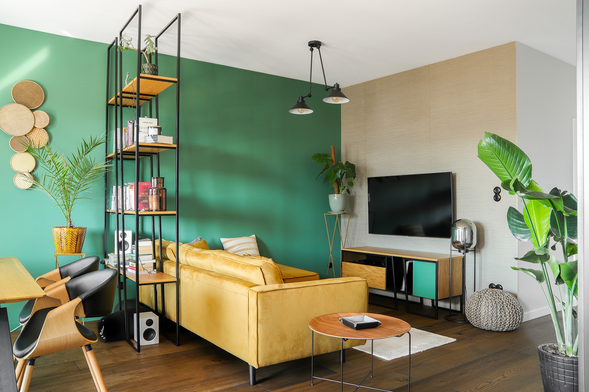 Nowoczesny salon z żółtą sofą, regałem z książkami i zieloną ścianą. Widoczne elementy dekoracyjne i oświetlenie, tworzące przytulną atmosferę wnętrza.