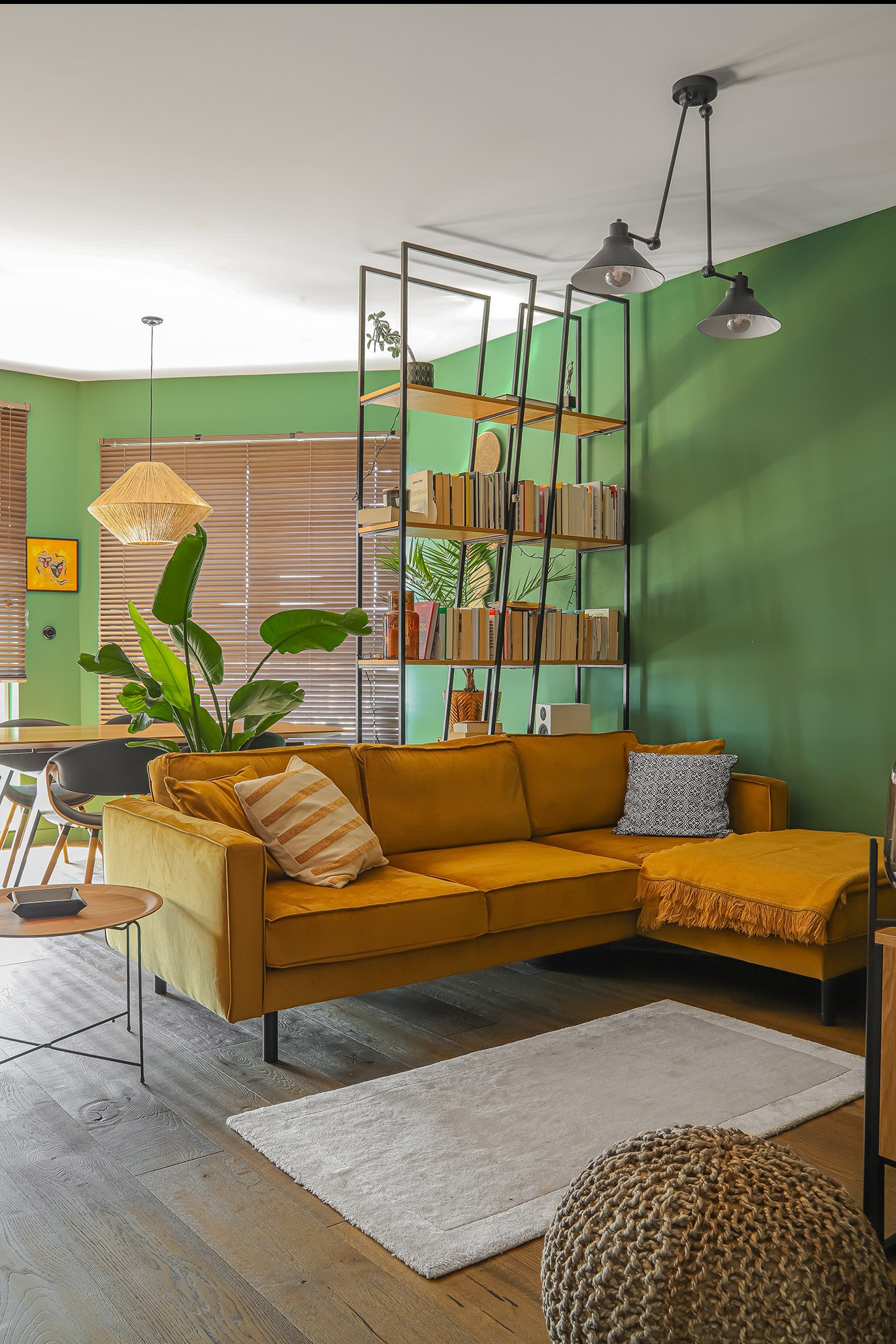 Stylowy salon z zielonymi ścianami, musztardową sofą narożną, regałem z książkami i drewnianą podłogą. Widoczne detale: lampy wiszące, dywan i rośliny doniczkowe.
